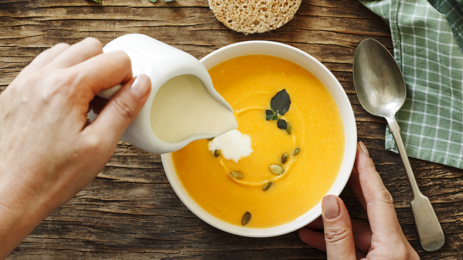 Hand gießt helle Flüssigkeit in orangefarbene Suppe.