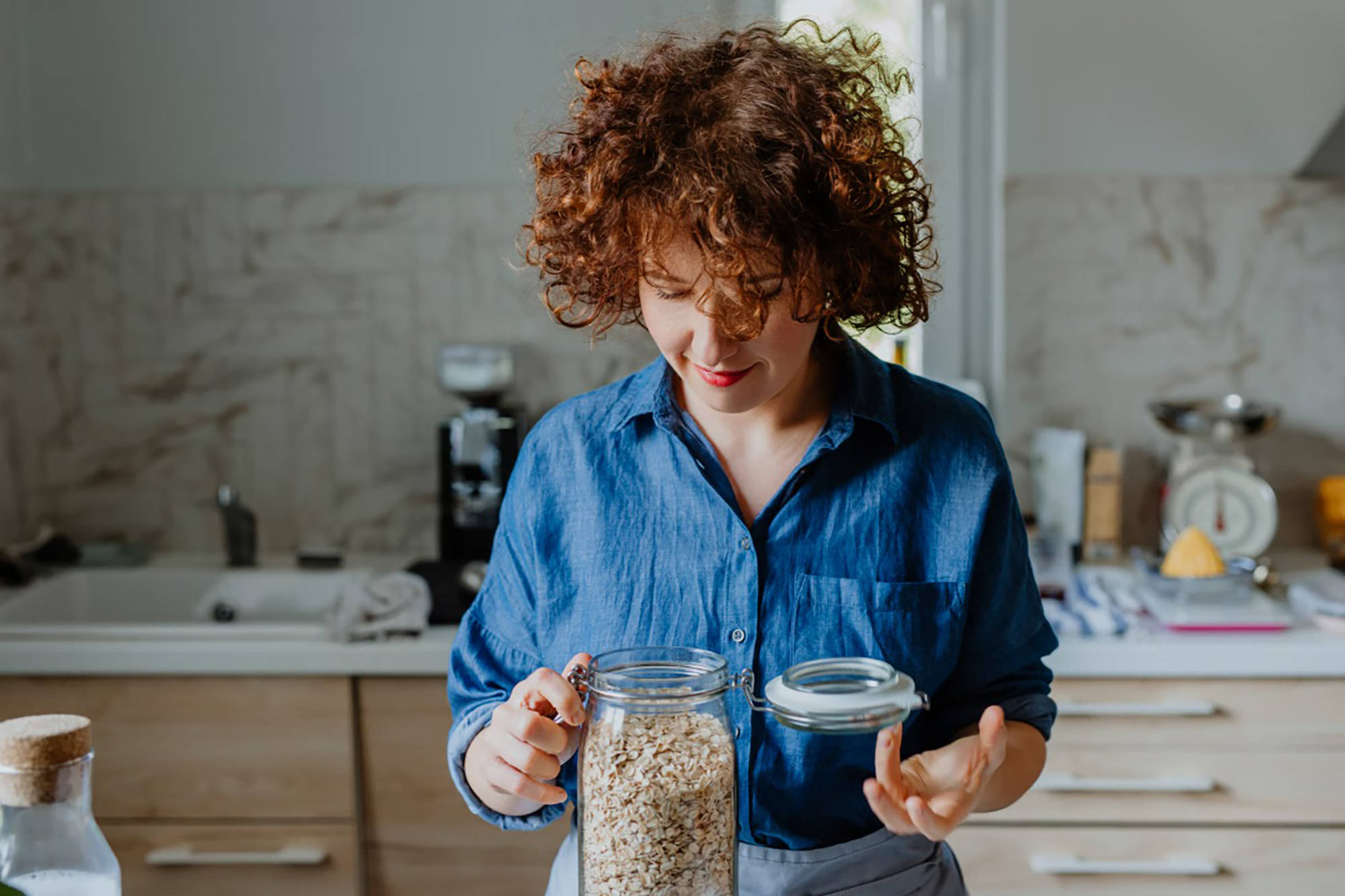 Frau in blauer Bluse öffnet ein Vorratsgals mit Haferflocken in der Küche.