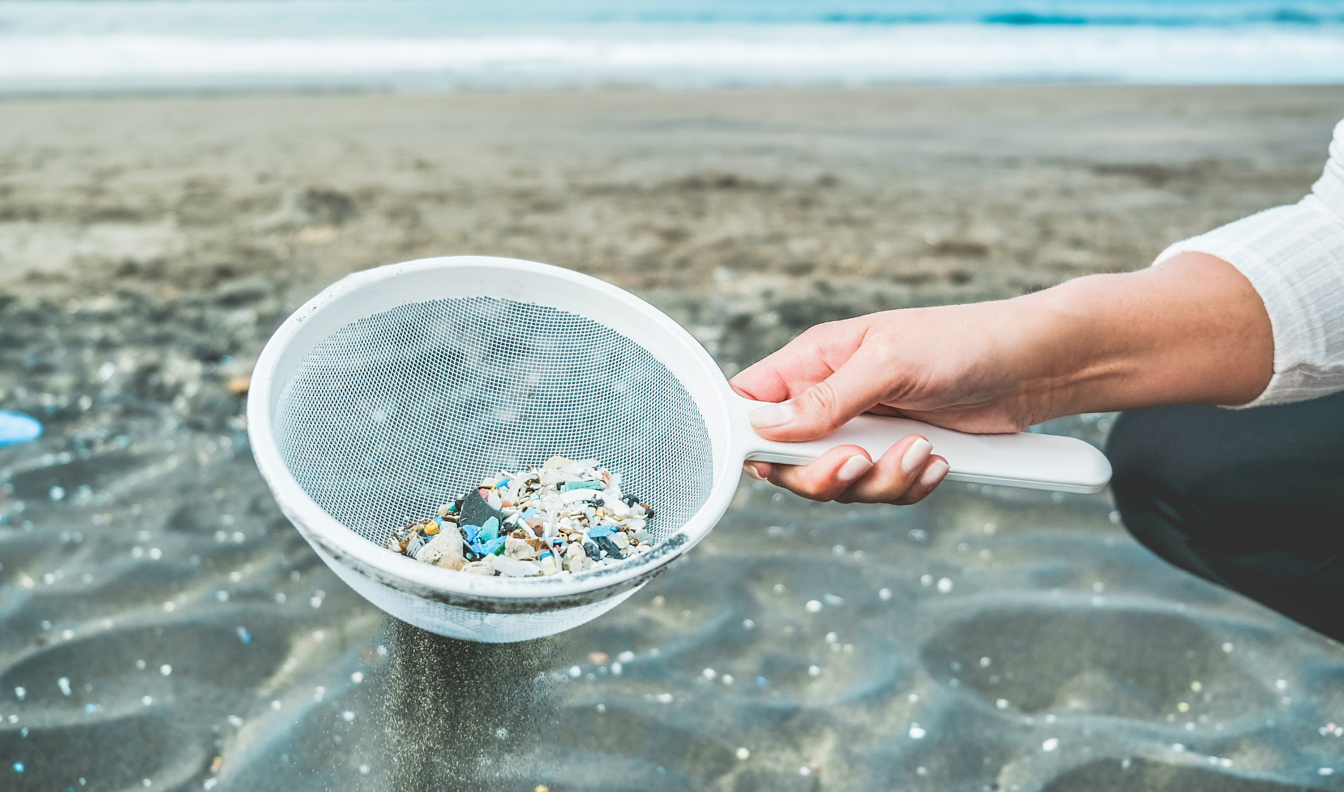 Hand mit Sieb voller Kunststoffteilchen am Strand.