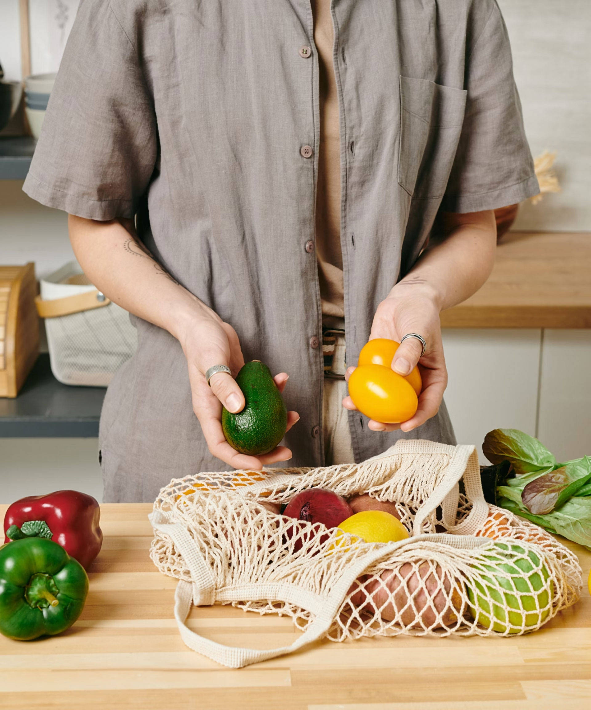 Person in einer Küche nimmt eine Avocado und gelbe Tomaten aus dem Netzbeutel, der mit frischem Gemüse gefüllt ist.