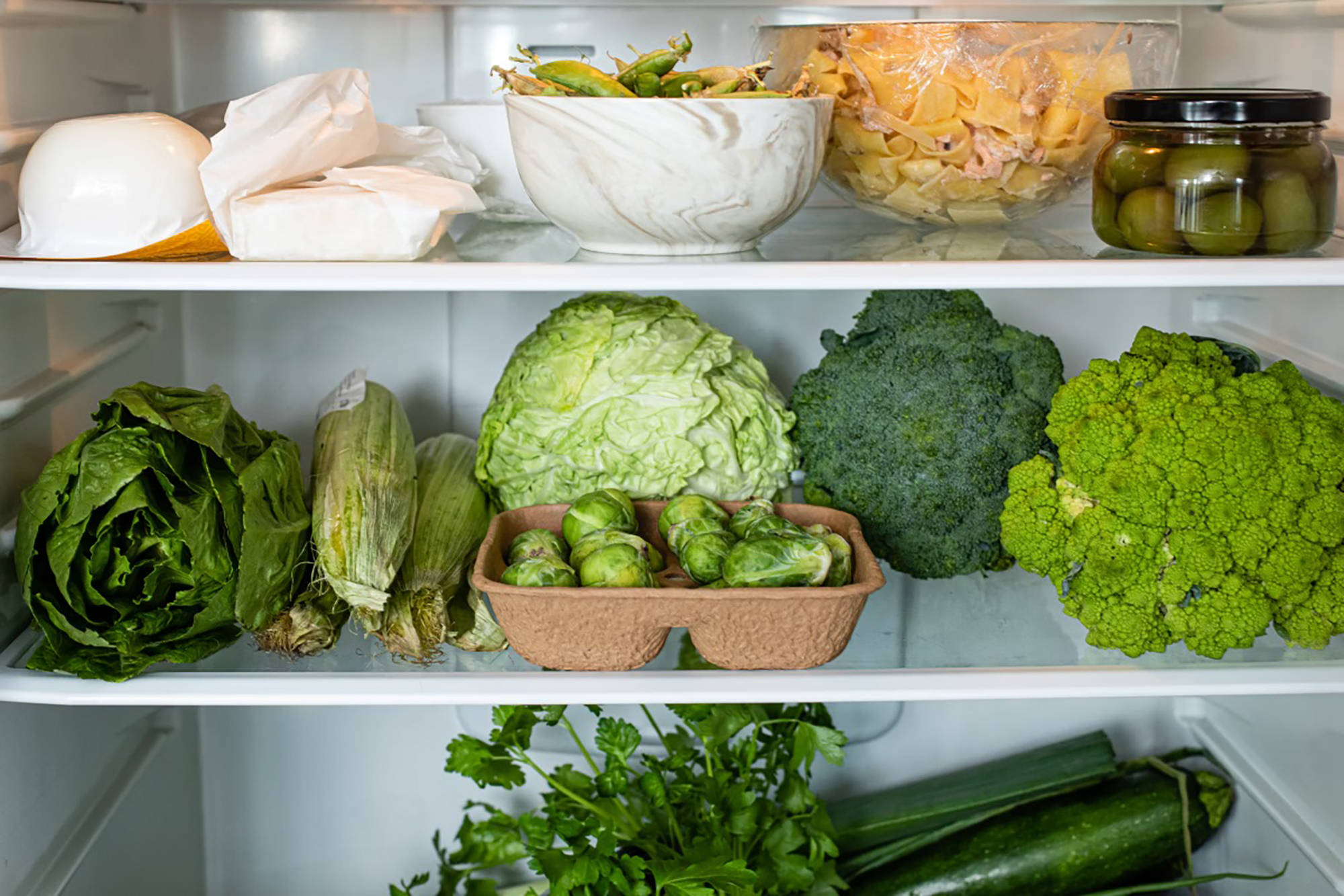Offener Kühlschrank, darin frisches grünes Gemüse, Käse und Oliven .
