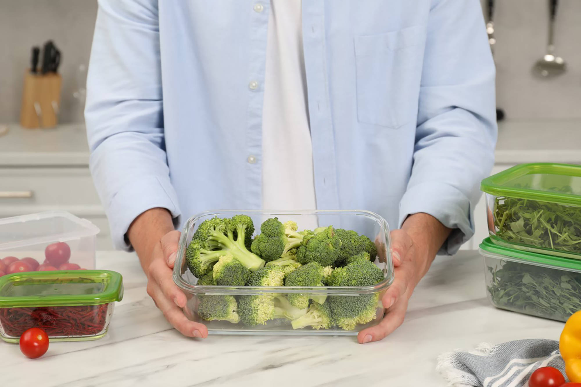 Person hält eine Frischhaltebox mit frischem Brokkoli, umgeben von weiteren Gemüsebehältern in der Küche.