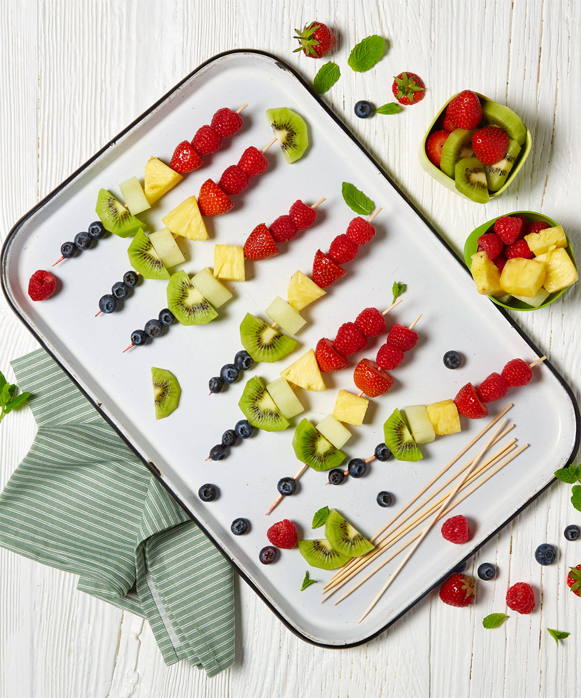 Bunte Obst-Spieße aus Kiwi, Ananas und Himbeeren liegen auf einem weißen Tablett.