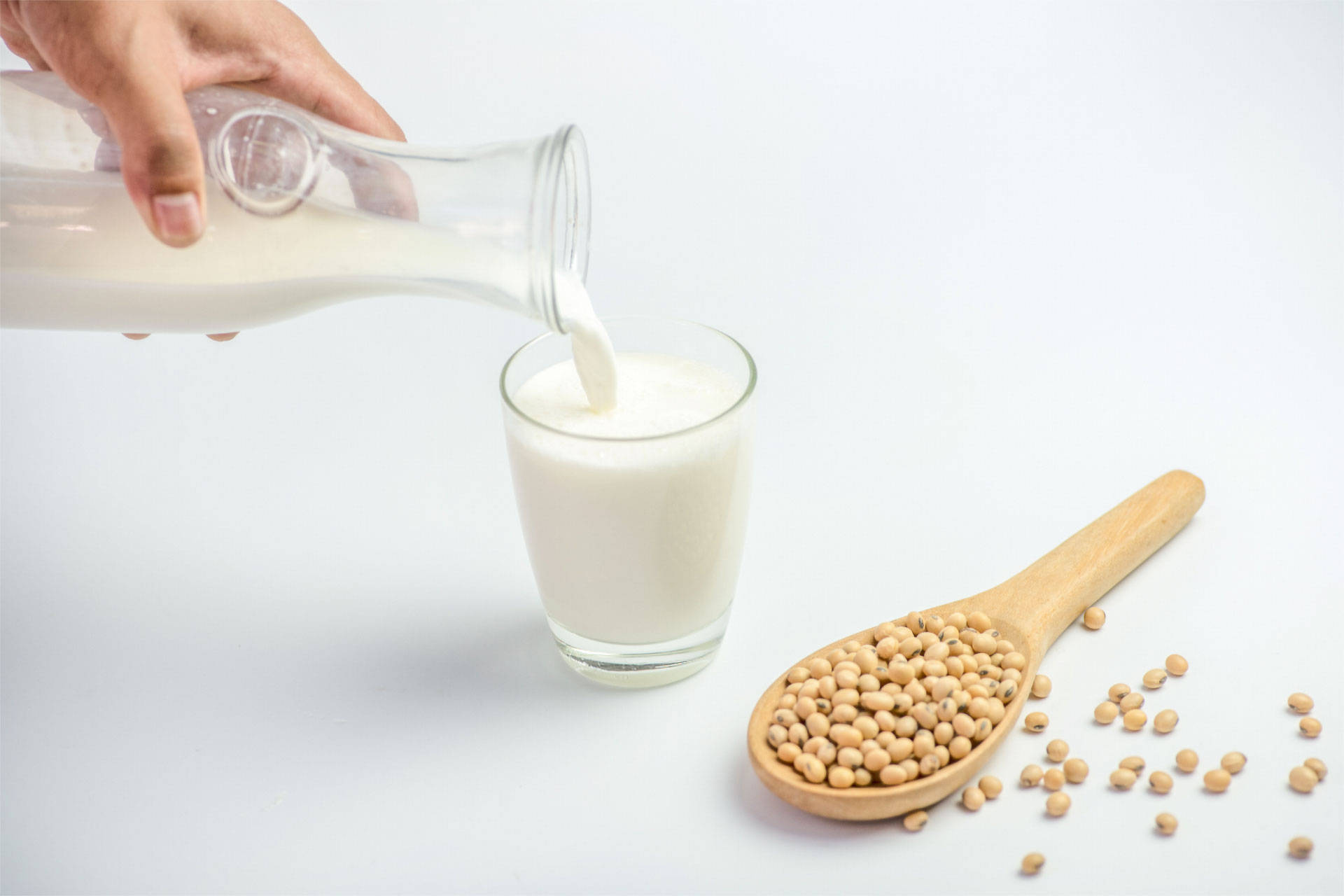 Jemand gießt Sojamilch aus einer Glaskaraffe in ein Glas, neben einem Holzlöffel mit Sojabohnen auf weißem Hintergrund.