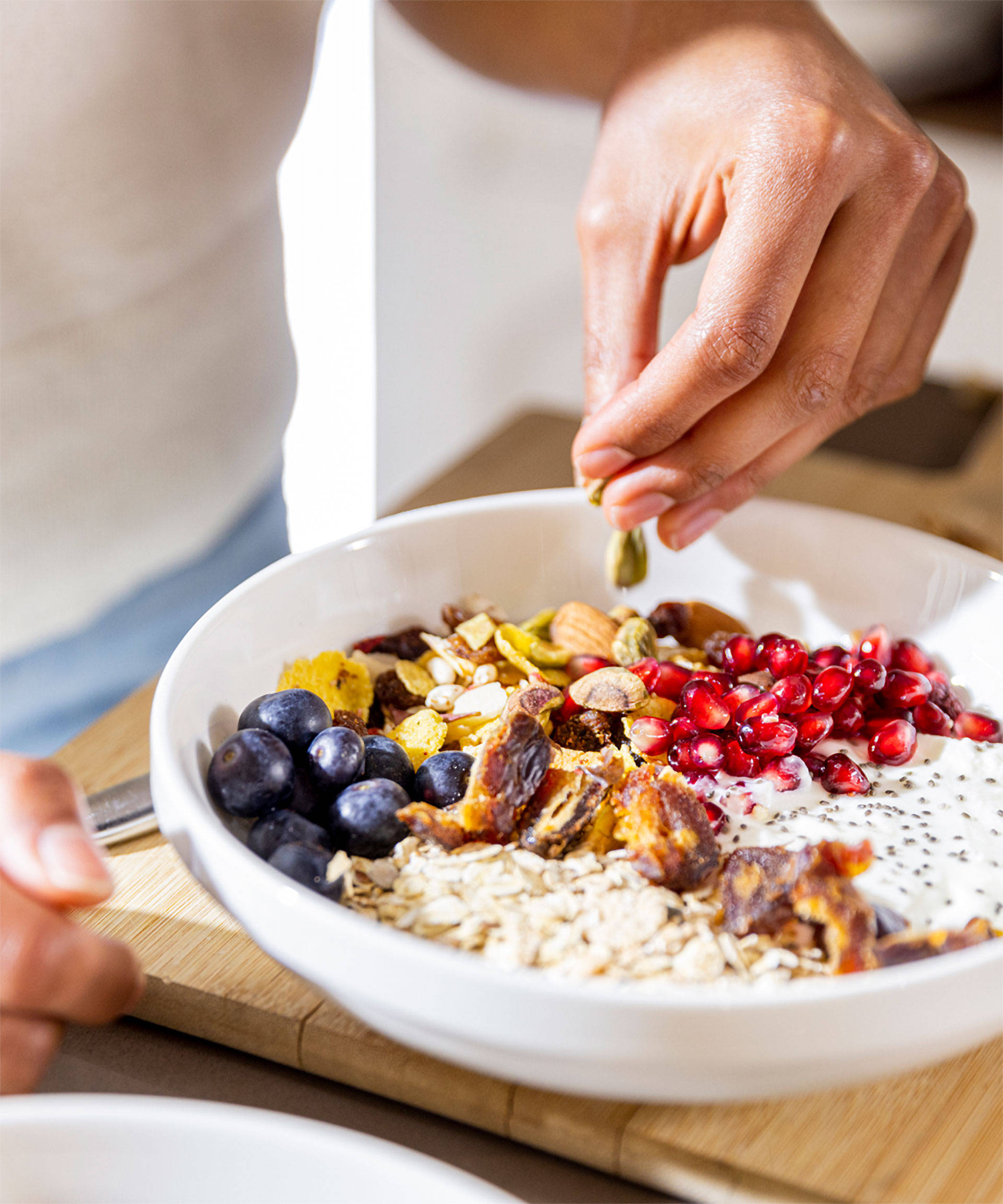 Person garniert eine Frühstücksschale mit Haferflocken, Blaubeeren, Granatapfelkernen, Nüssen und Joghurt auf einem Holz