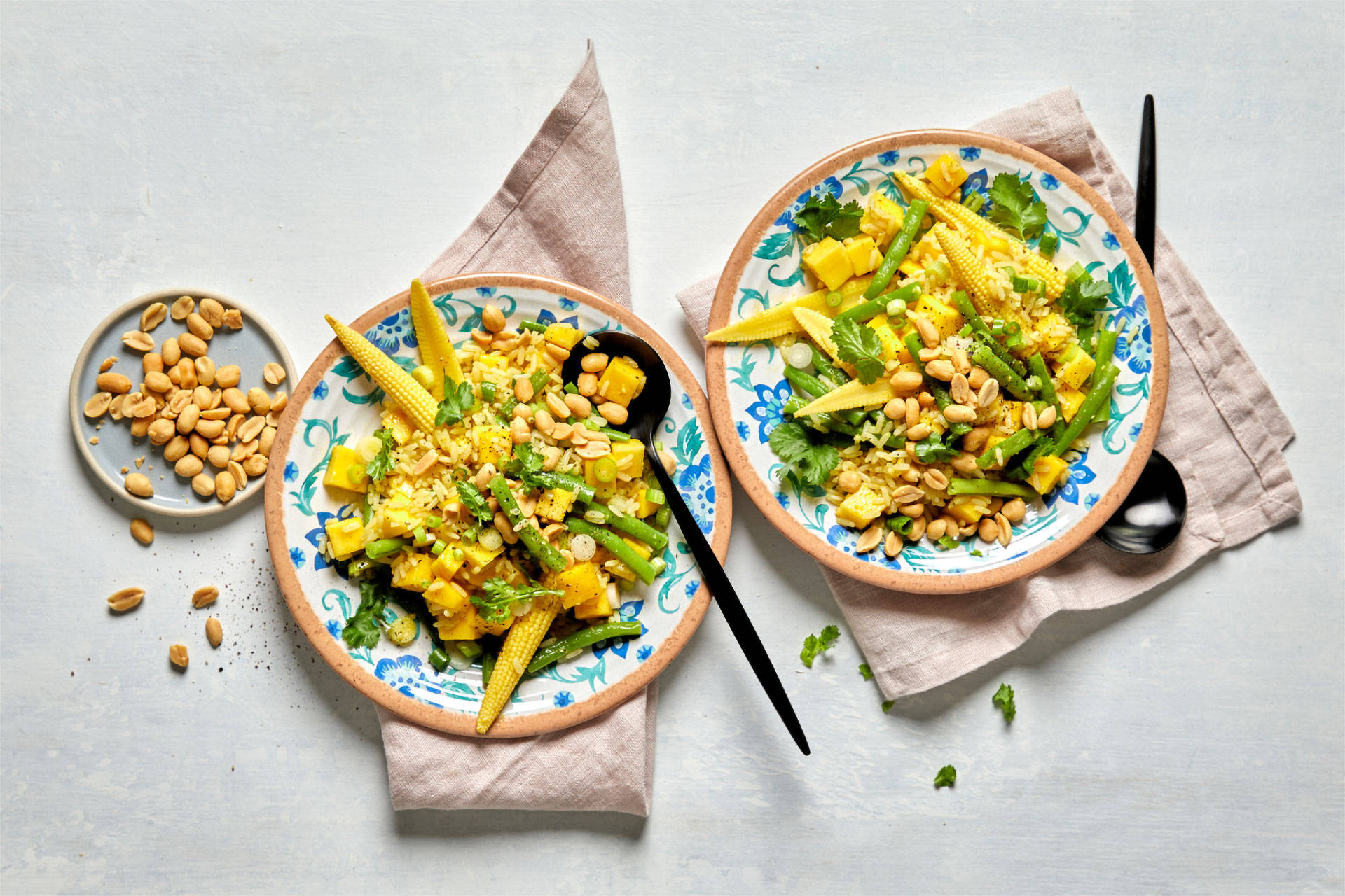 Reissalat mit Babymais, grünen Bohnen, Mango und Erdnüssen, serviert in zwei Schalen mit frischen Kräutern und Löffeln.