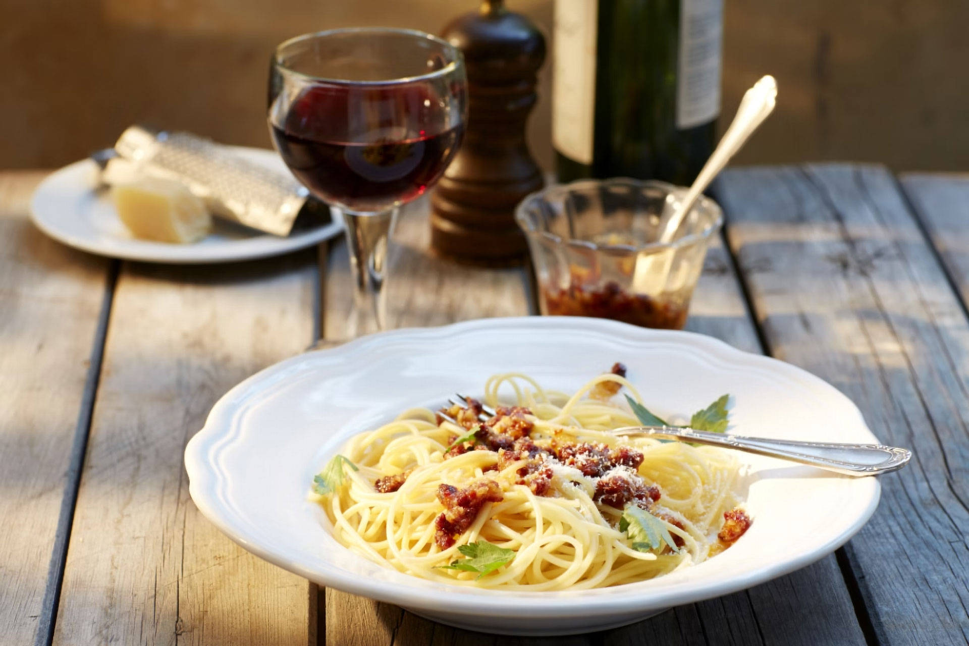 Holztisch mit Spaghetti-Teller und gefülltem Rotweinglas, dahinter unscharf Rotweinflasche, Gewürzmühle und Glasschale.