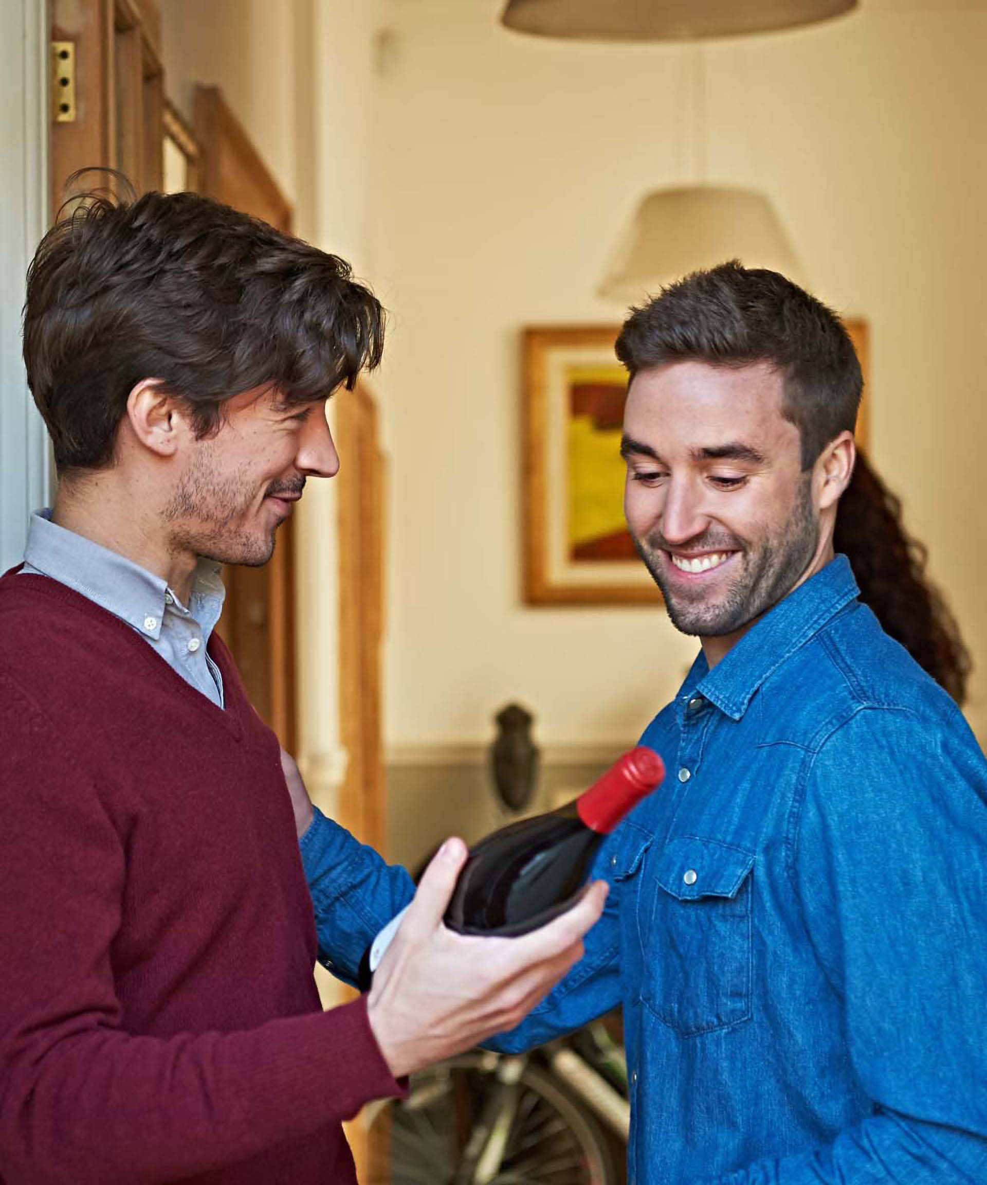 Ein Mann übereicht einem anderen Mann eine Flasche Rotwein einer Eingangstür.