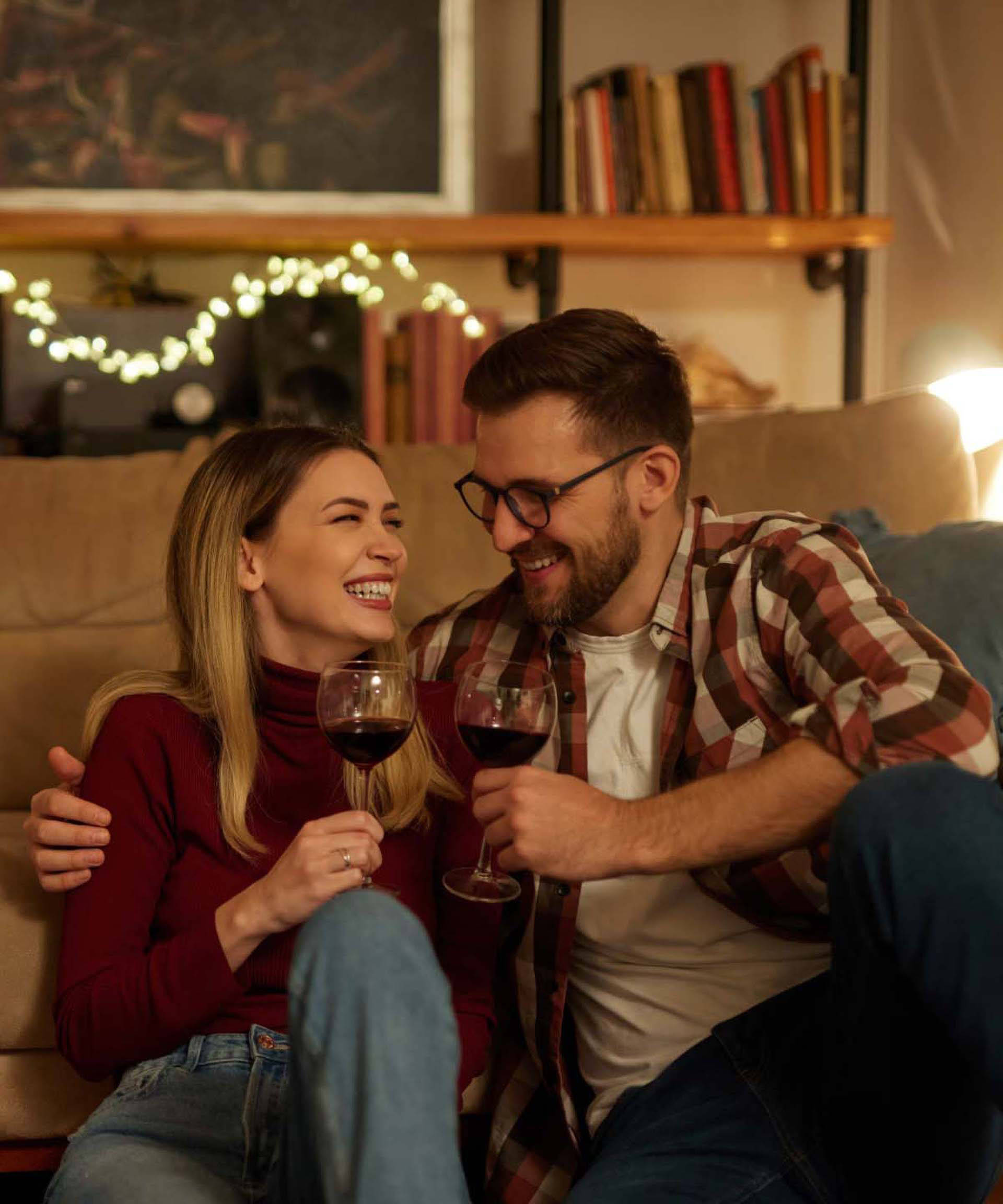 Ein Paar sitzt lachend auf dem Boden vor einem Sofa in einem gemütlichen Wohnzimmer mit gedimmtem Licht. Sie halten Rotw