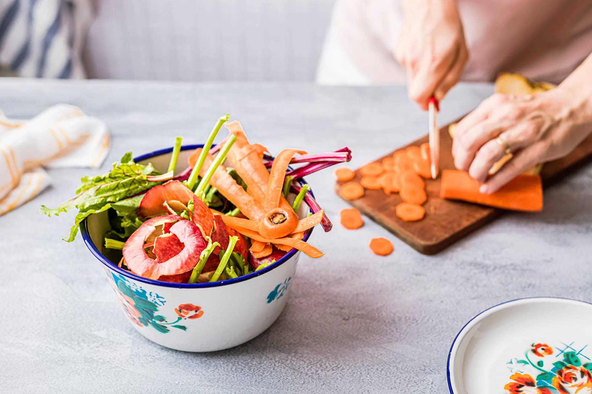 Person schneidet Karotten auf Holzbrett, Gemüsereste in bunter Schüssel im Vordergrund.