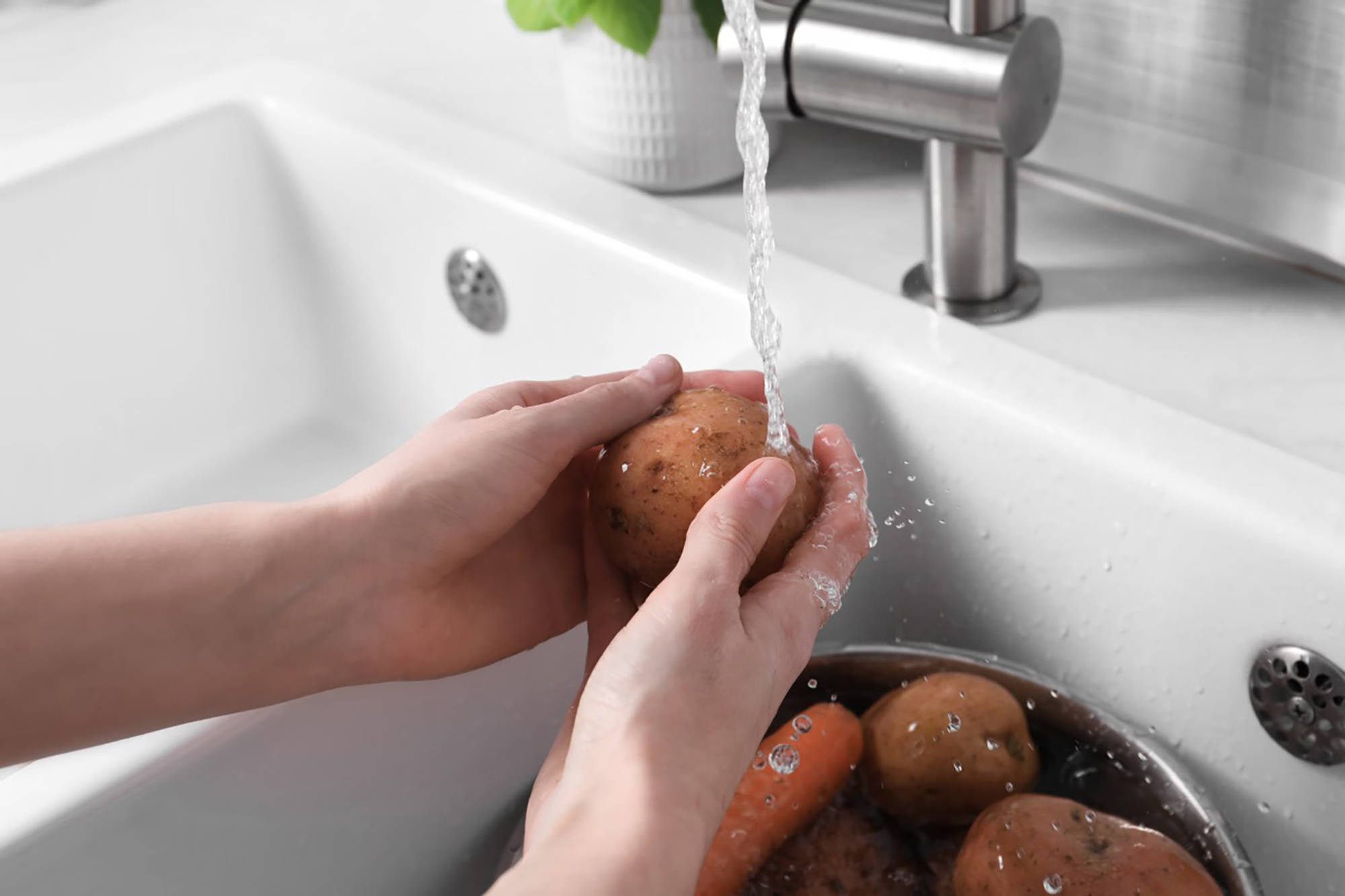 Jemand wäscht Kartoffeln unter fließendem Wasser in weißem Spülbecken ab.