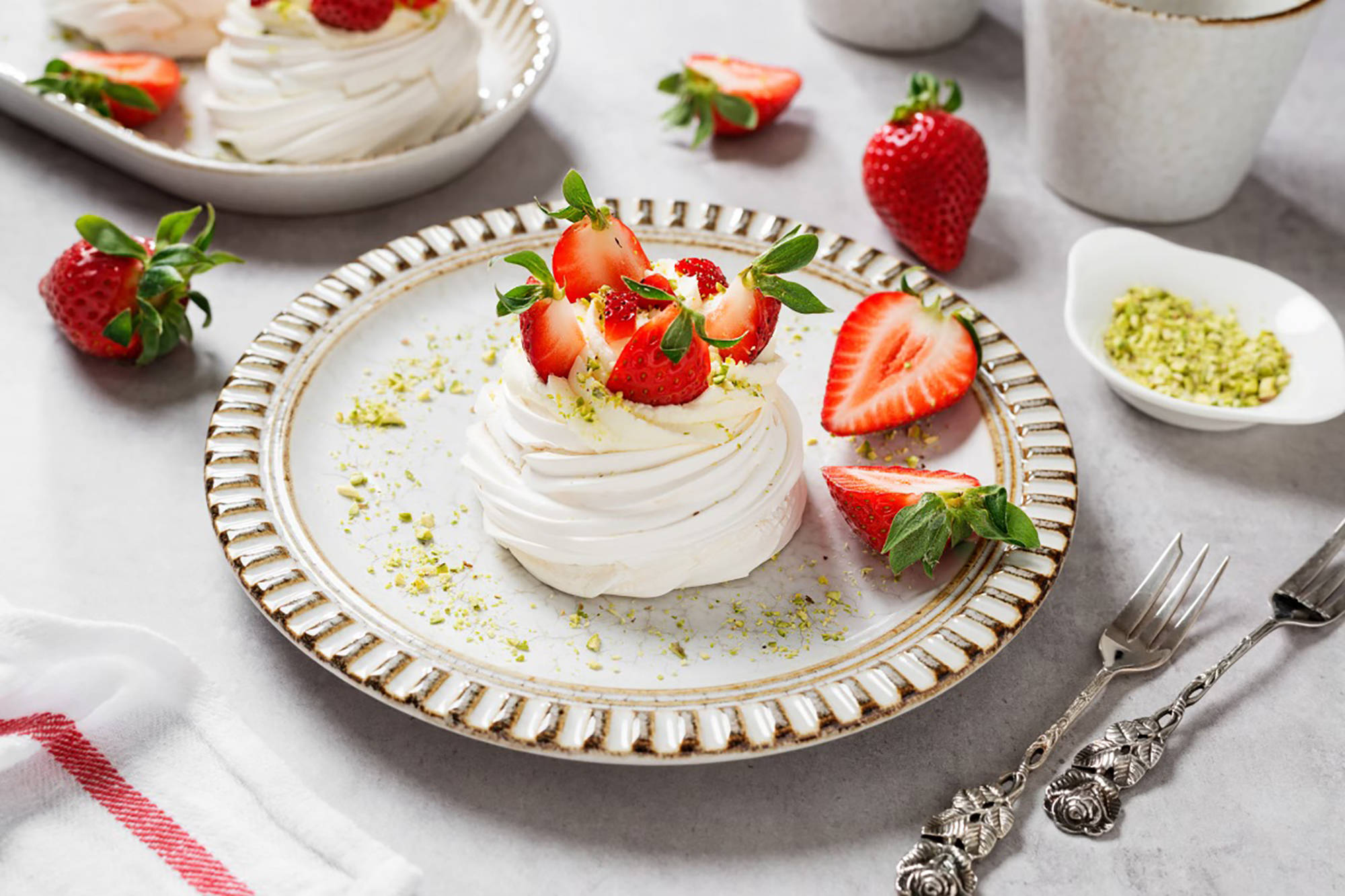 Pavlova-Dessert mit frischen Erdbeeren und Pistazien auf elegantem Teller, umgeben von weiteren Erdbeeren und Besteck.