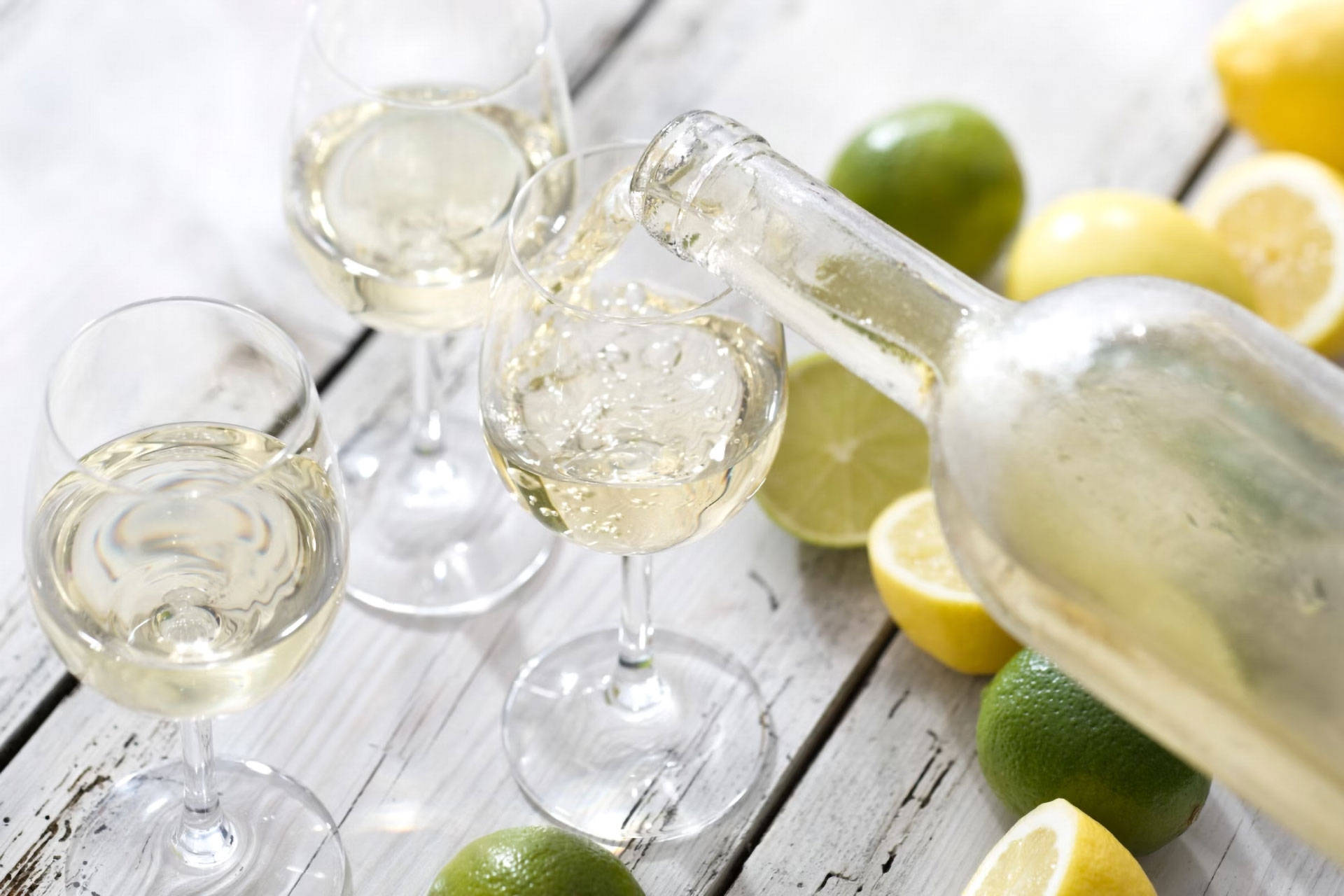 Auf einem Tisch stehen drei Weingläßer, die gerade mit Riesling befüllt werden, daneben halbierte Limetten und Zitronen.