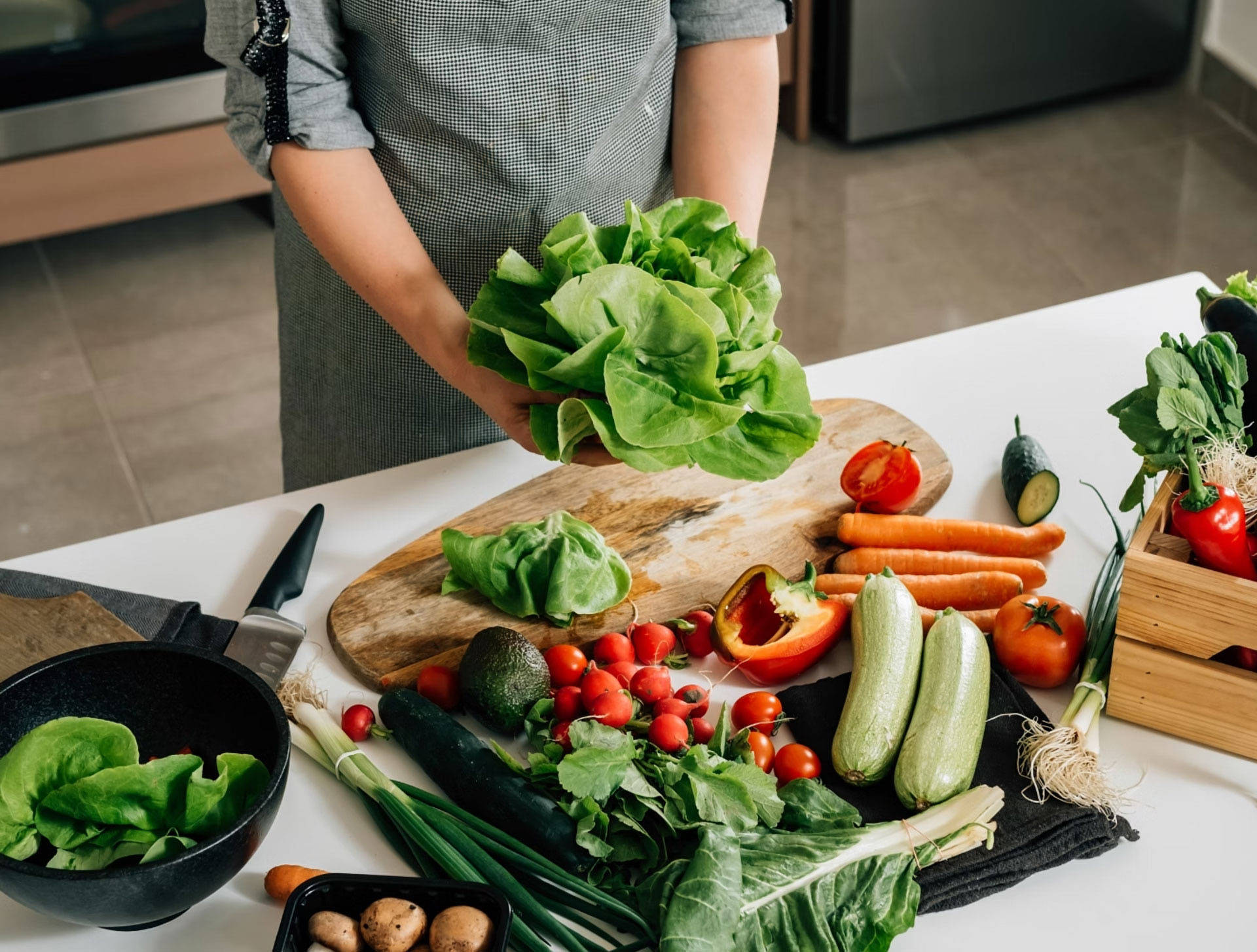Person bereitet buntes Saisongemüse, wie Salat, Paprika, Gurken, Radieschen und Avocado auf einem Küchentisch vor.