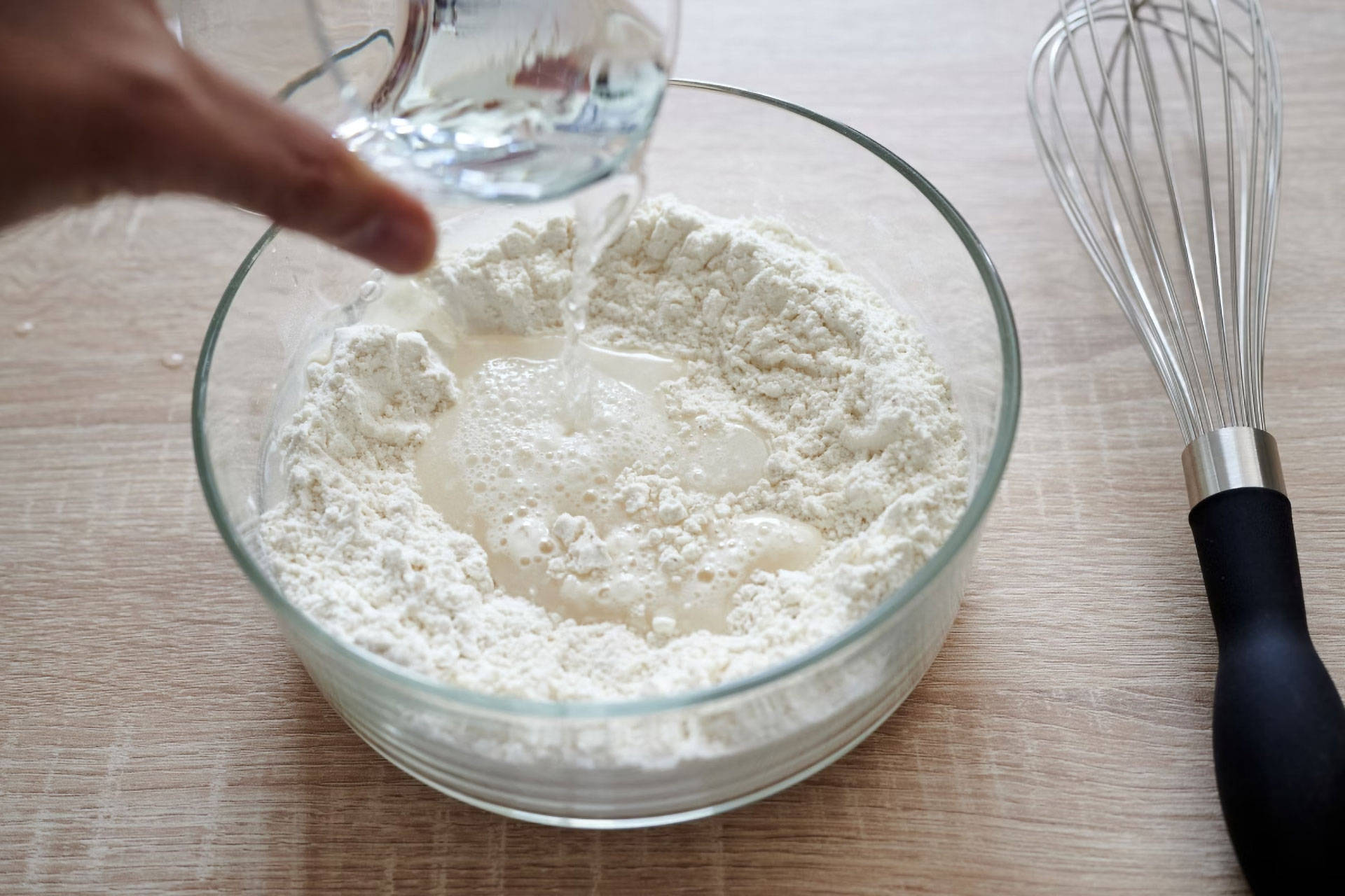 Eine Person gibt Wasser in eine Schüssel Mehl. Daneben liegt ein Schneebesen zum Umrühren.