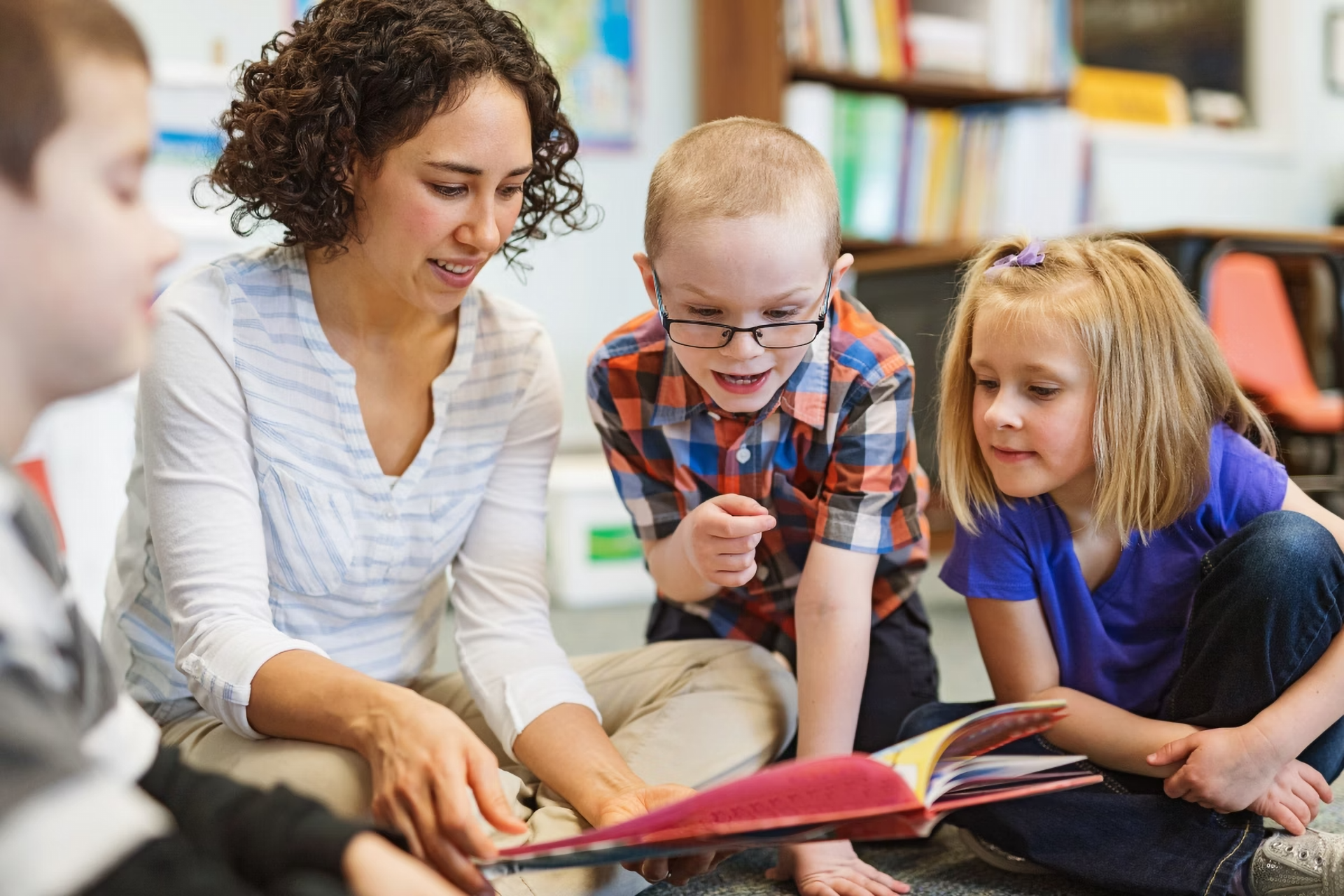 Eine Frau liest drei Kindern etwas aus einem Buch vor.