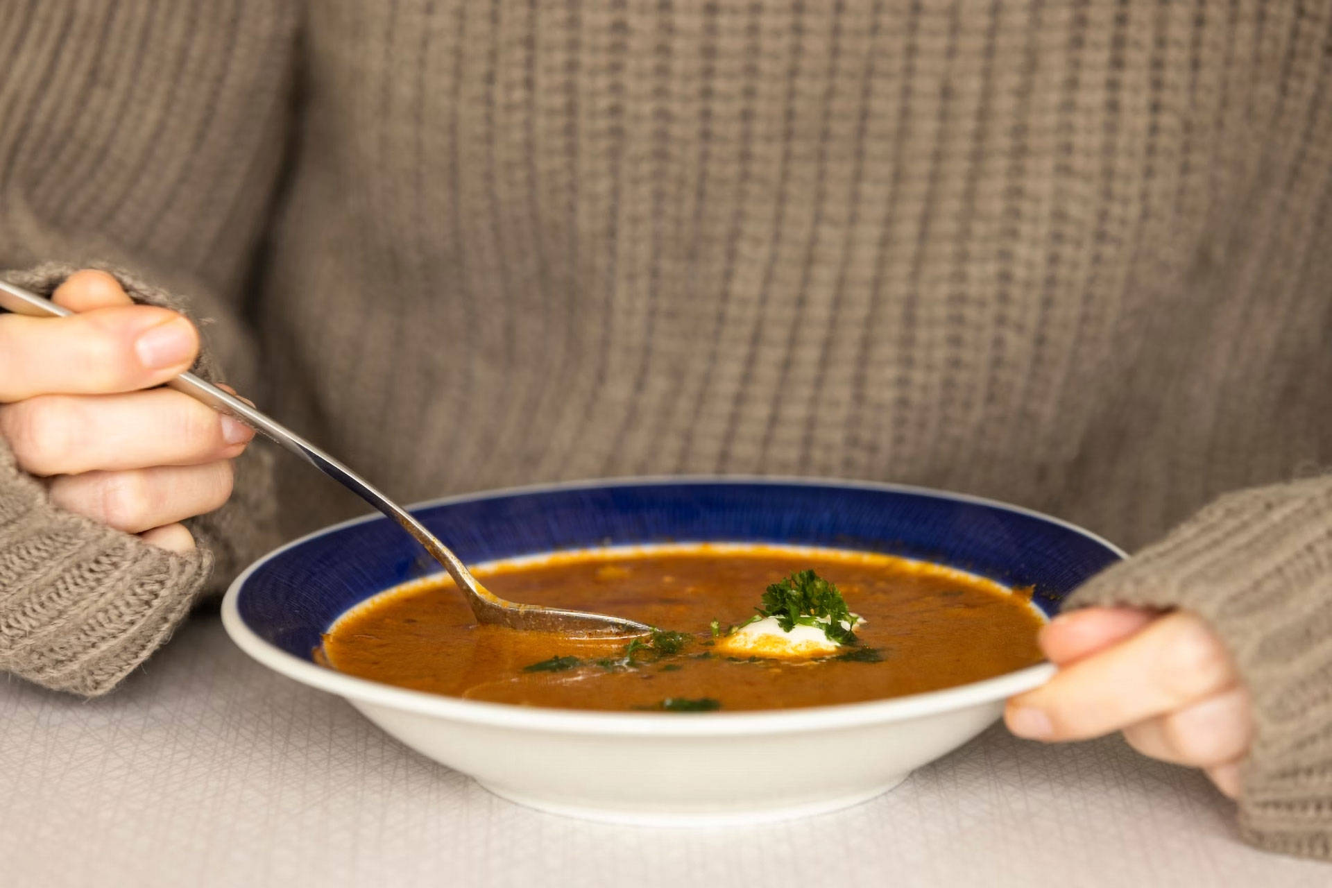 Person hält einen Löffel in einen Teller mit heißer Suppe, garniert mit Sahne und Kräutern.