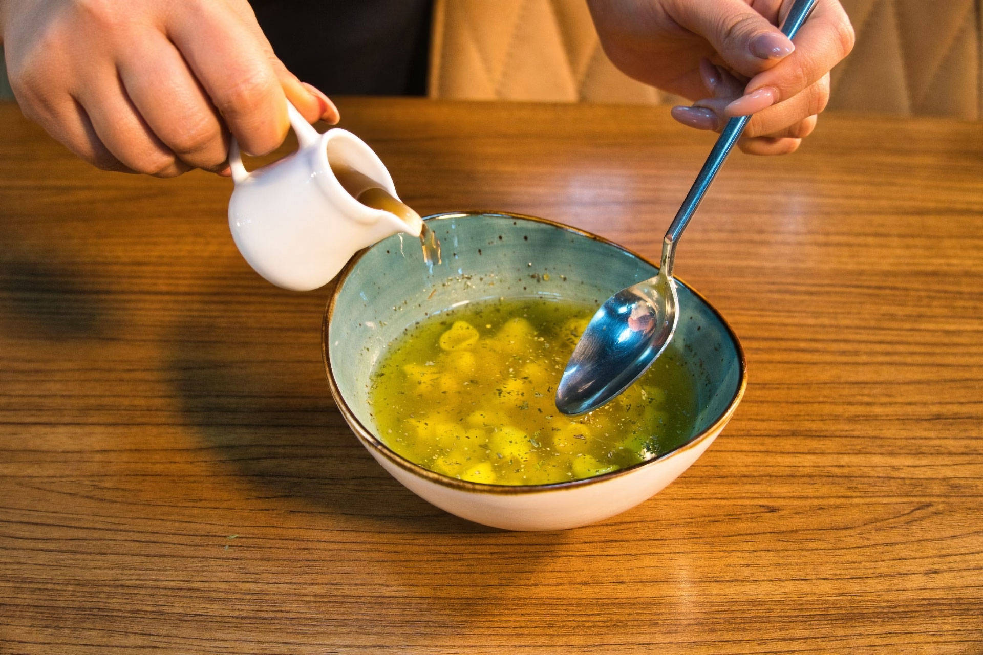 Person gießt Soße aus einer kleinen Kanne in eine Schüssel mit Suppe, hält dabei einen Löffel in der anderen Hand.