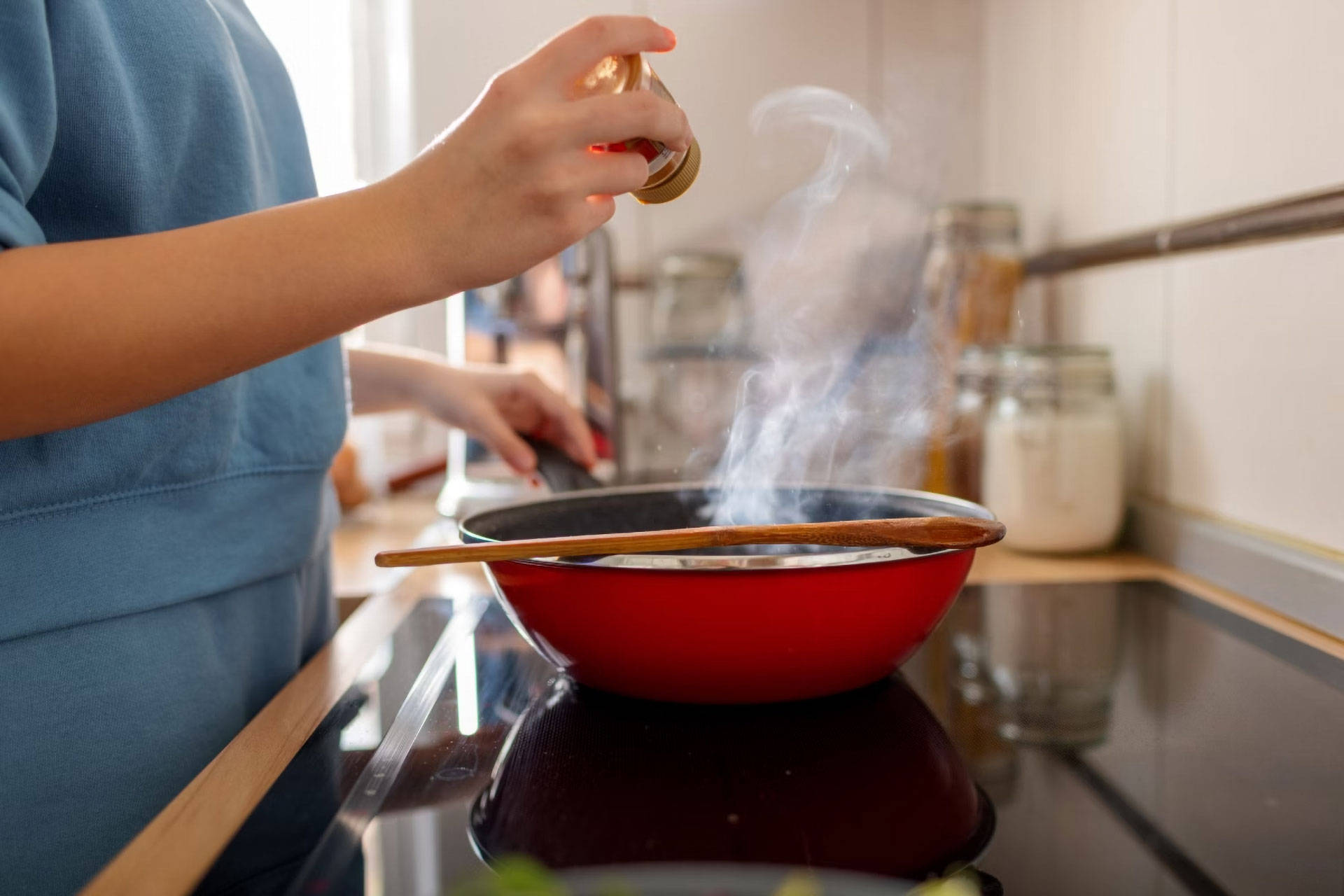 Person würzt ein Gericht in einer dampfenden roten Pfanne auf dem Herd. Holzlöffel liegt auf dem Rand.