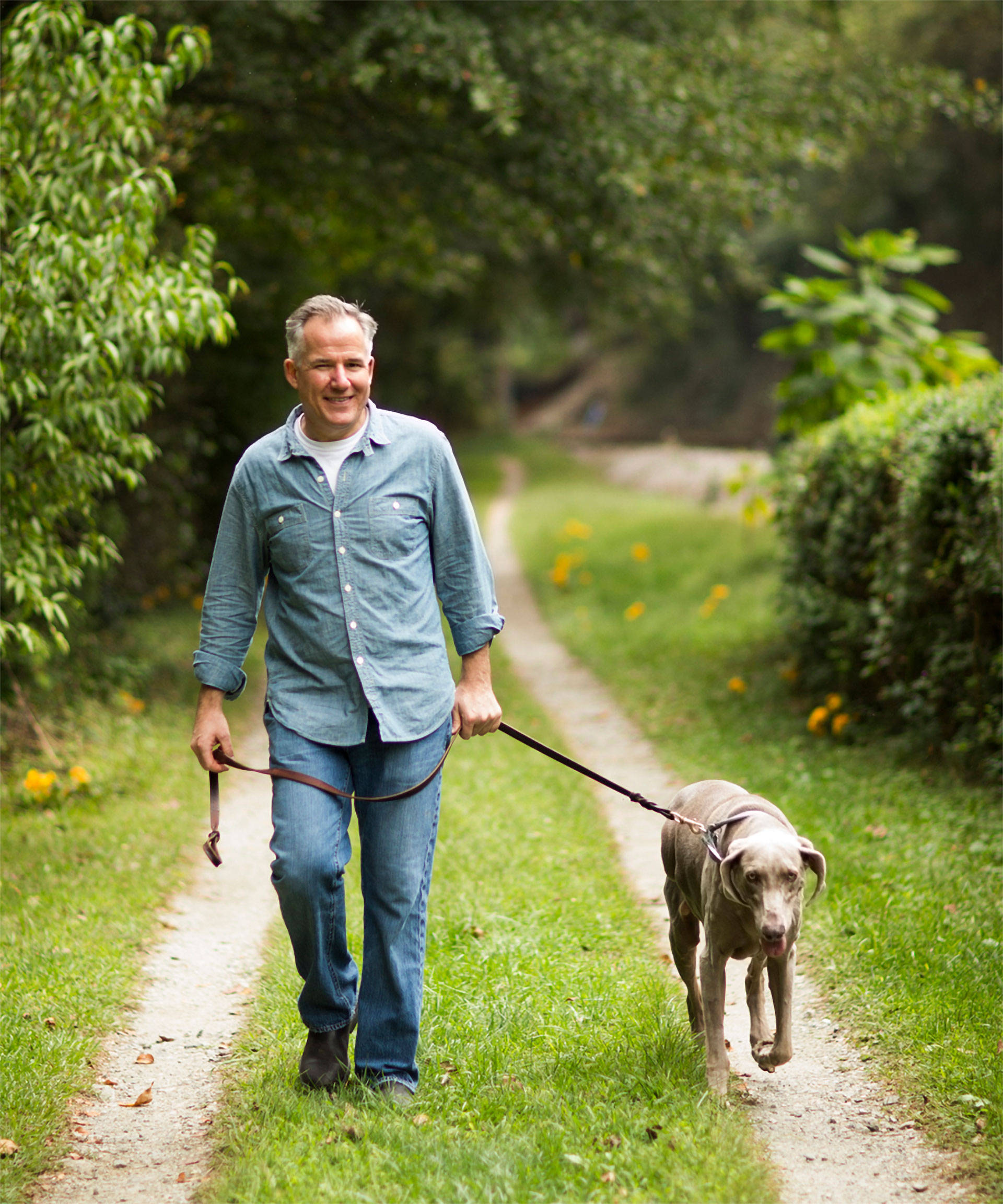 Mann geht lächelnd mit seinem Hund an der Leine auf einem Waldweg spazieren.
