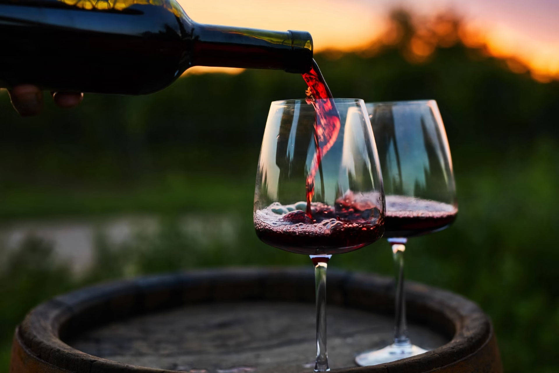 Zwei Weingläser auf einem hölzernen Weinfass werden gerade mit Rotwein gefüllt, im Hintergrund unscharf Natur und Sonnen