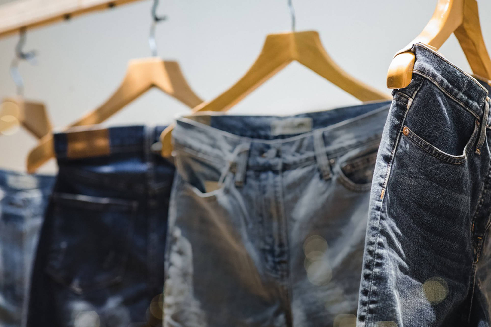  Drei Jeans in verschiedenen Blautönen, laserwashed, auf Holzkleiderbügeln in einem Laden hängend.