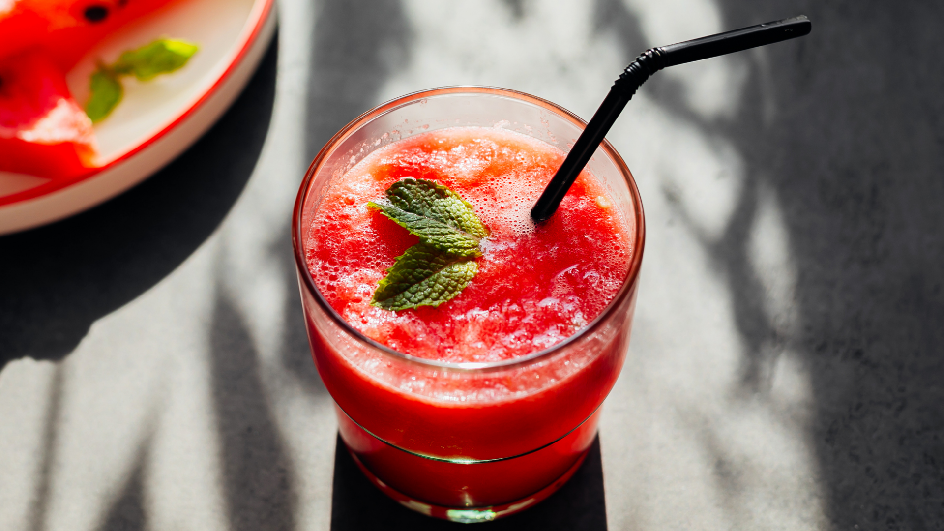 Wassermelonengetränk mit Minze und schwarzem Trinkhalm, Teller im Hintergrund.