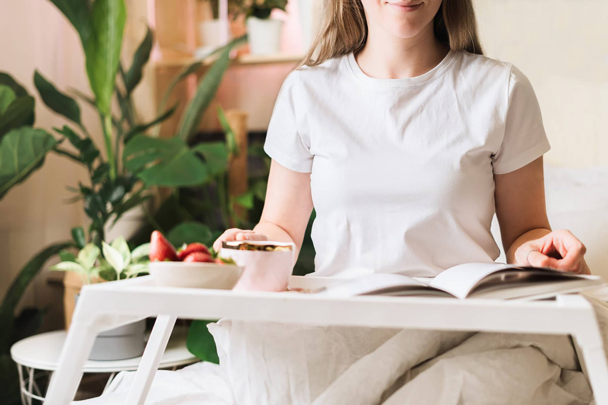 Frau sitzt im Bett mit einem Tablett und blättert durch ein Fotobuch. Auf dem Tablett stehen Erdbeeren und ein Getränk.