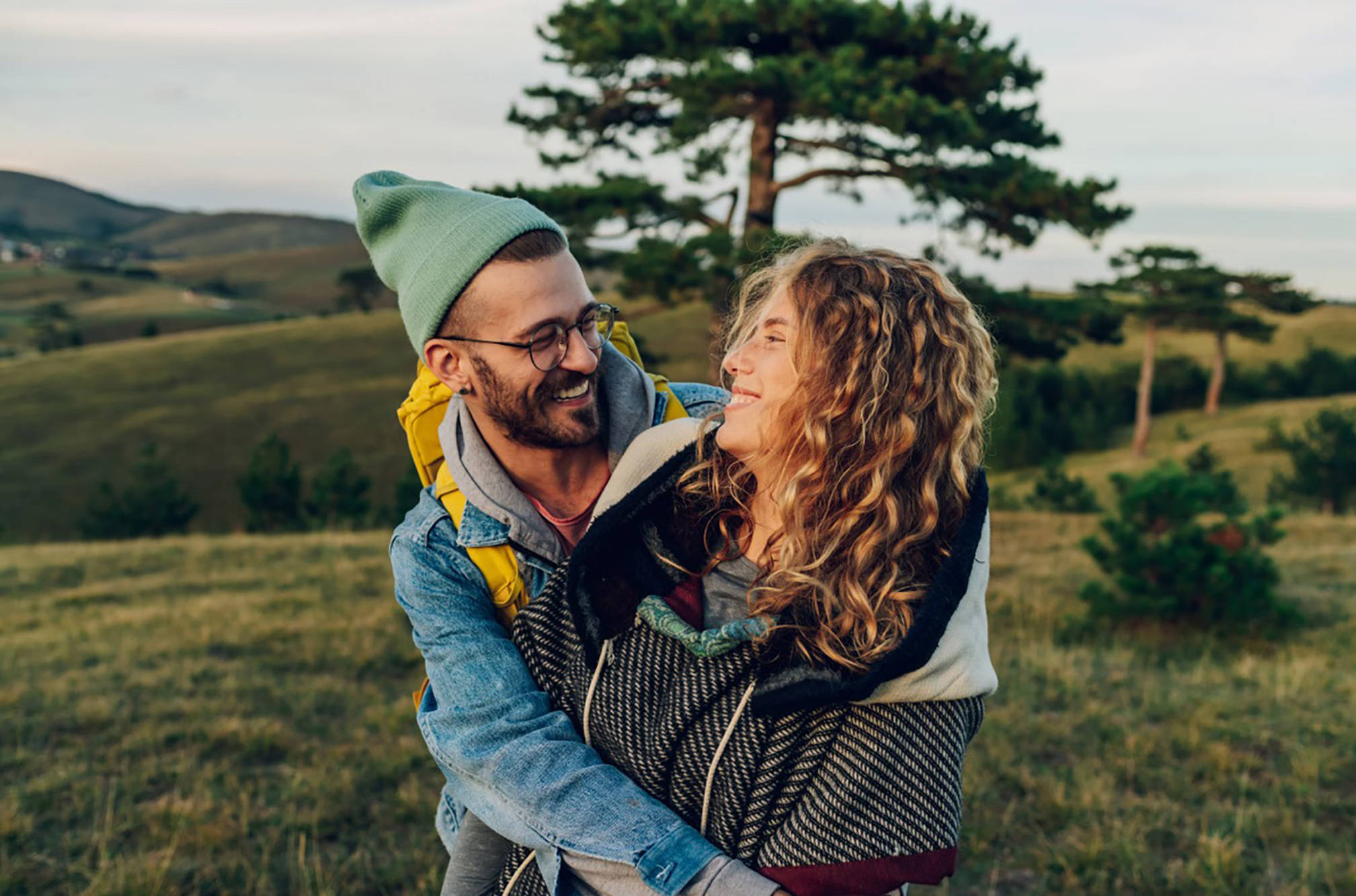 Lächelndes Paar in Outdoor-Kleidung umarmt sich auf einer Wiese. Hinter ihnen sind Bäume und grüne Hügel.