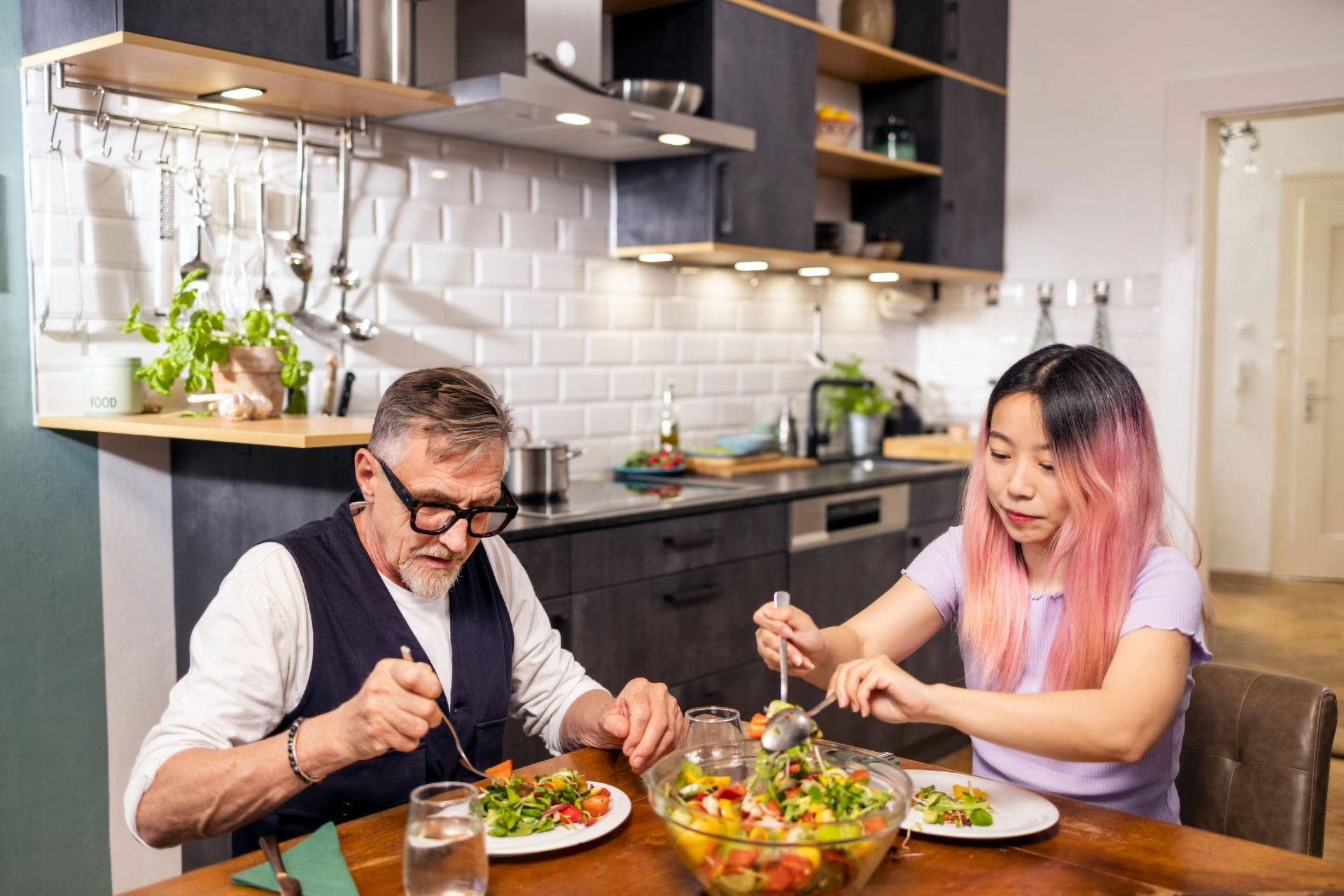Ein älterer Mann und eine junge Frau essen gemeinsam bunten Salat an einem Holztisch.
