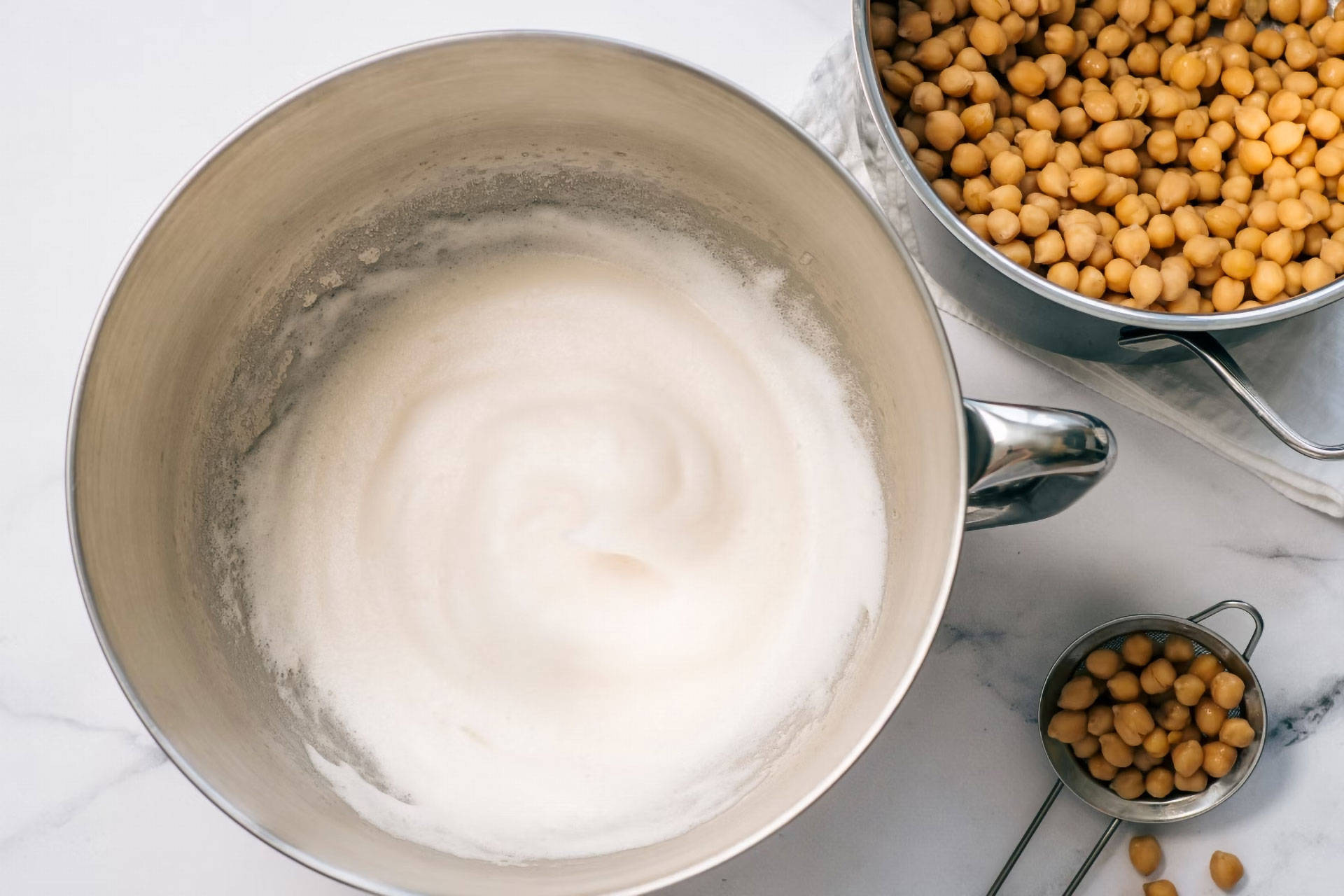 Ein Metallbecher, gefüllt mit geschlagener Aquafaba, daneben ein Sieb mit Kichererbsen.