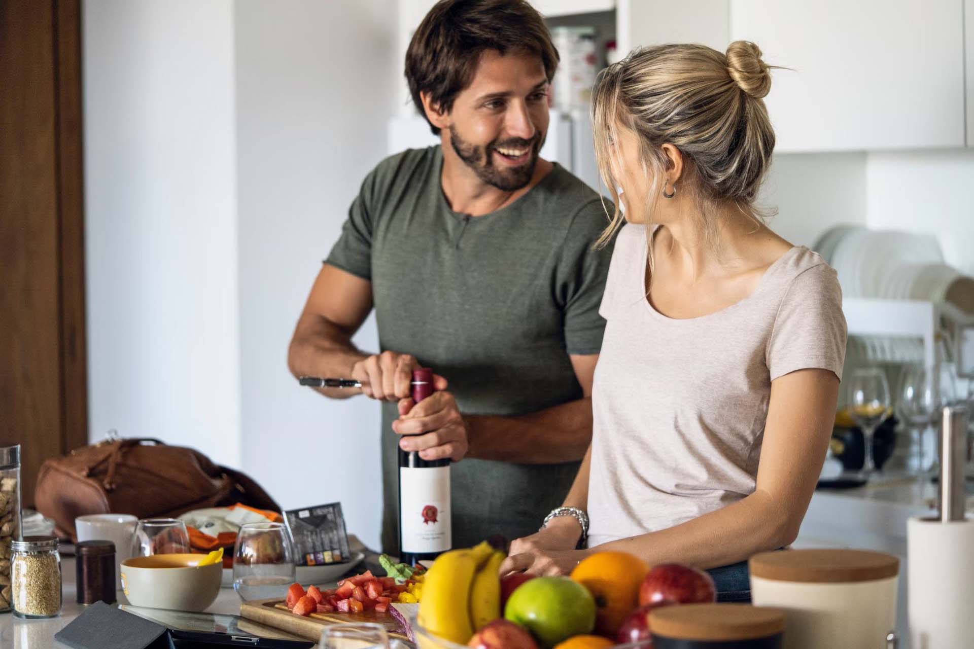 Ein Paar öffnet während des gemeinsamen Kochens lachend eine Flasche Rotwein.