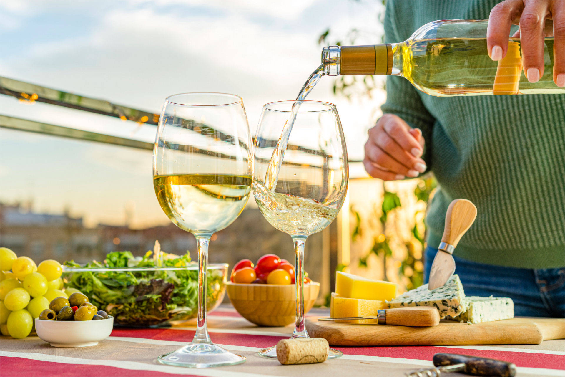 Person schenkt Weißwein in zwei Gläser auf einem Tisch mit Käse, Trauben und Salat bei Sonnenuntergang ein.