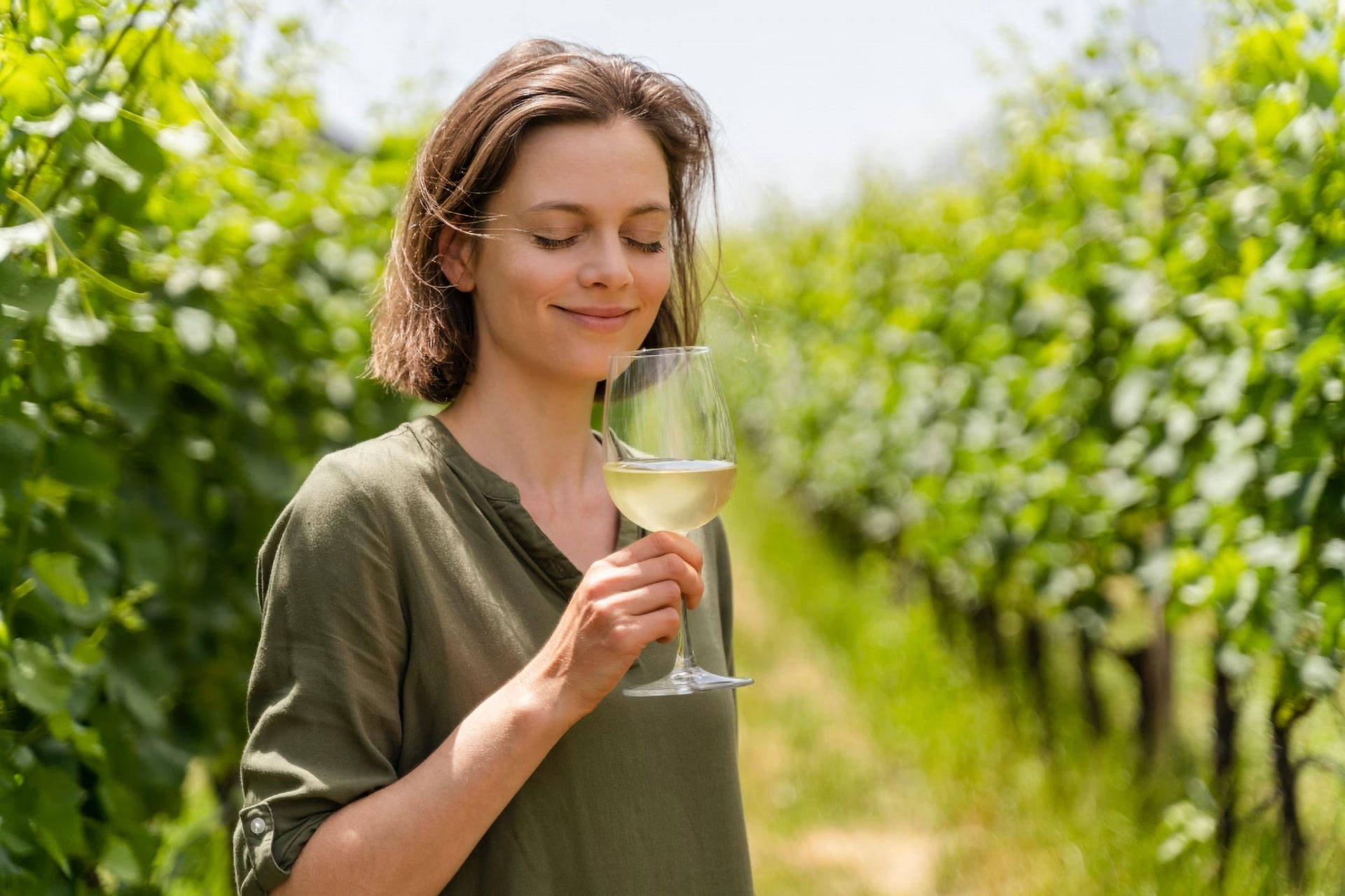 Eine Frau steht zwischen Weinreben mit einem Glas Weißwein in der Hand.