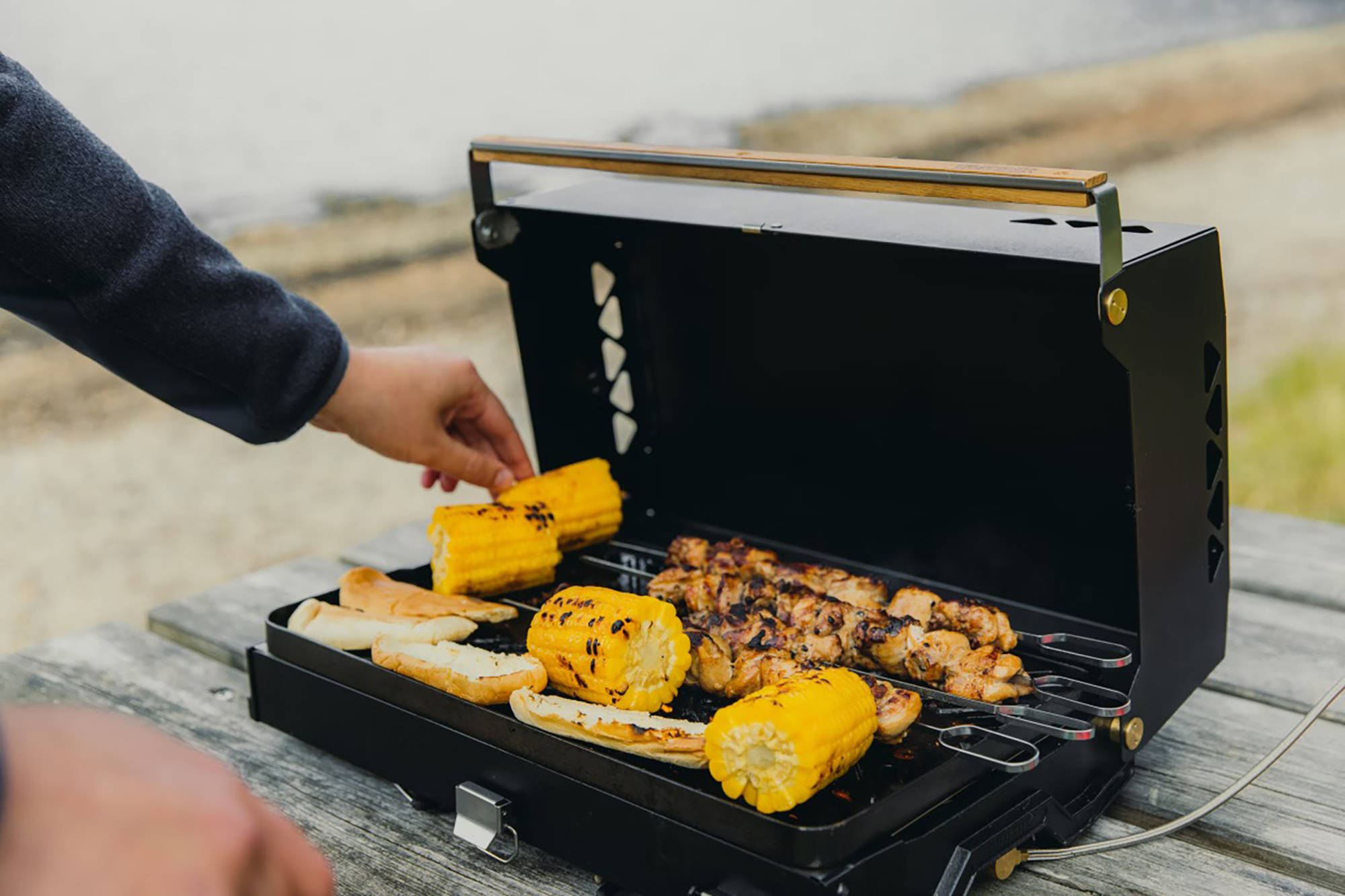 Person grillt Maiskolben und Fleisch auf einem tragbaren Grill, der im Freien auf einem Holztisch steht.