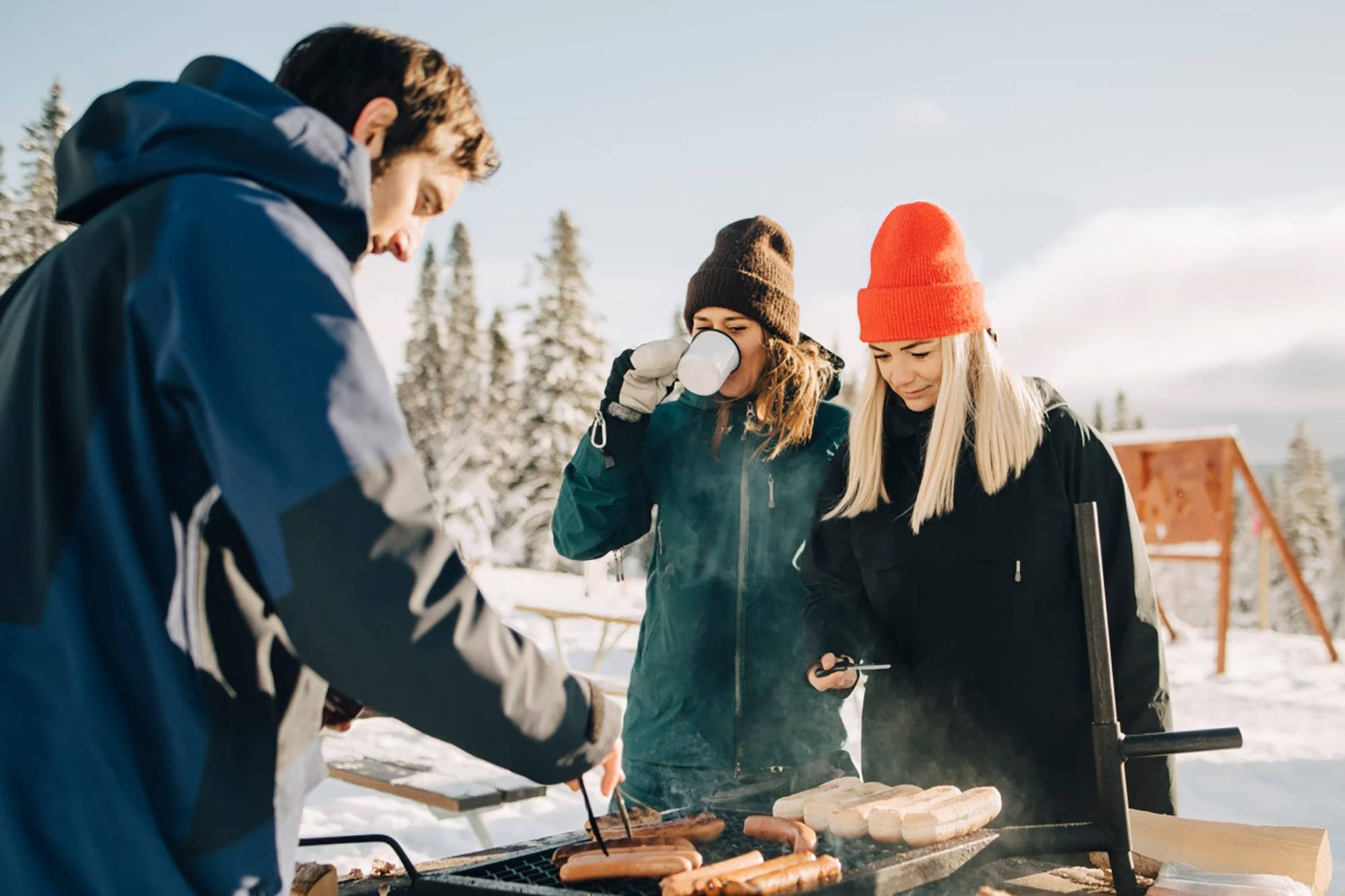 Drei Personen grillen Würste draußen im Winter auf einem Outdoor-Grill im Schnee.