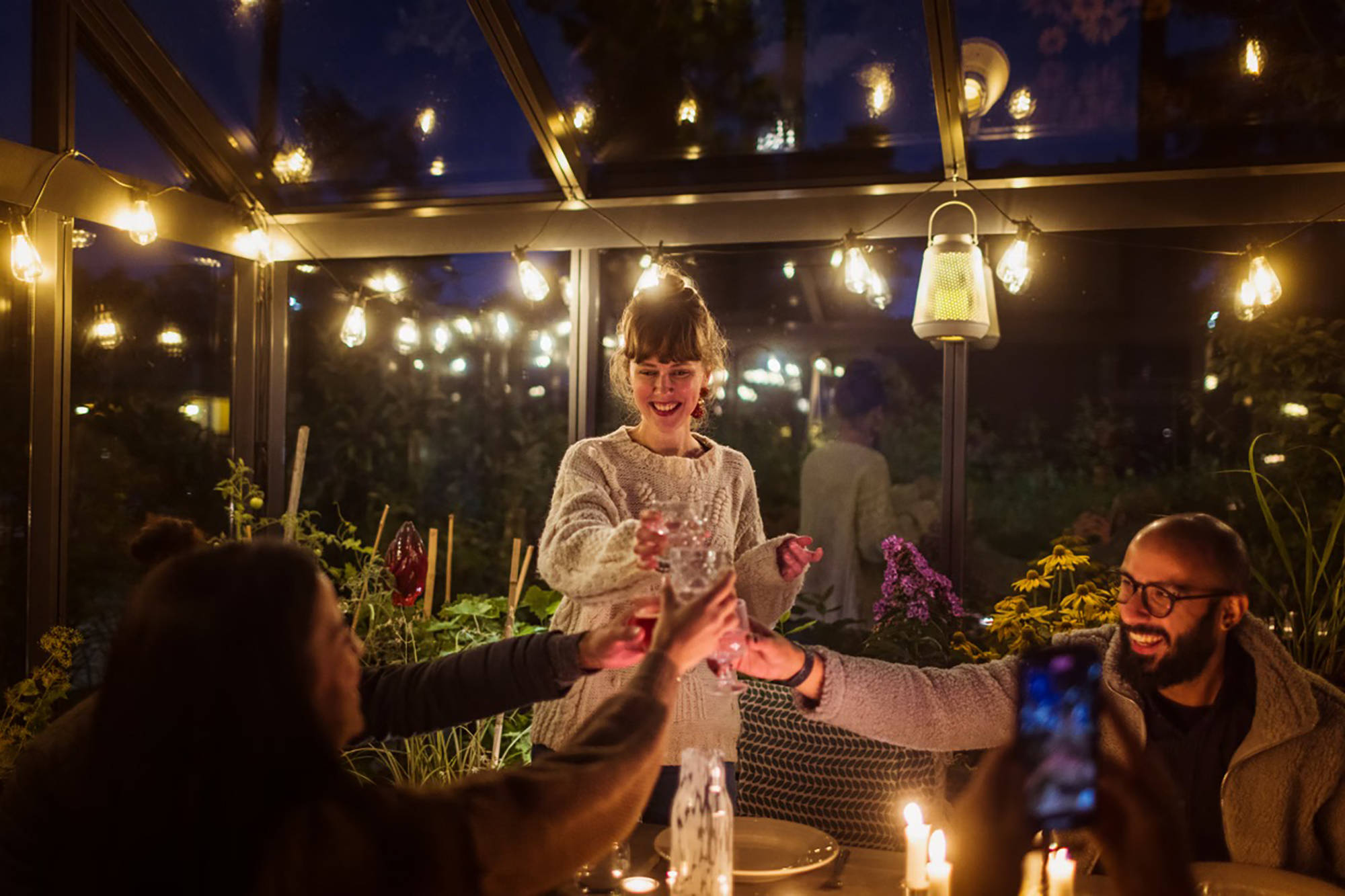 3 Personen stoßen in einem gemütlich beleuchteten Wintergarten mit Gläsern an.