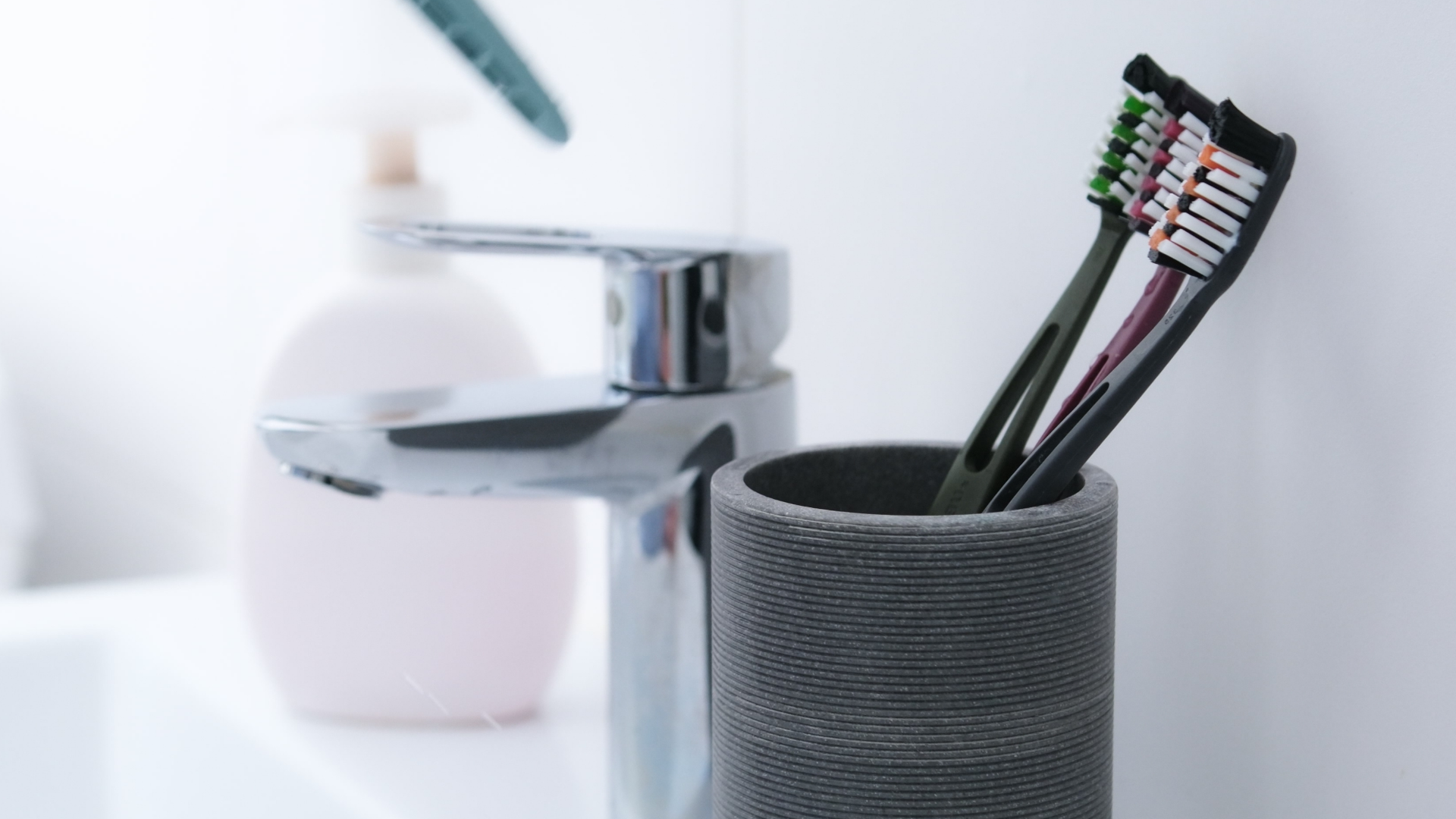 Drei Zahnbürsten im Becher neben Wasserhahn.