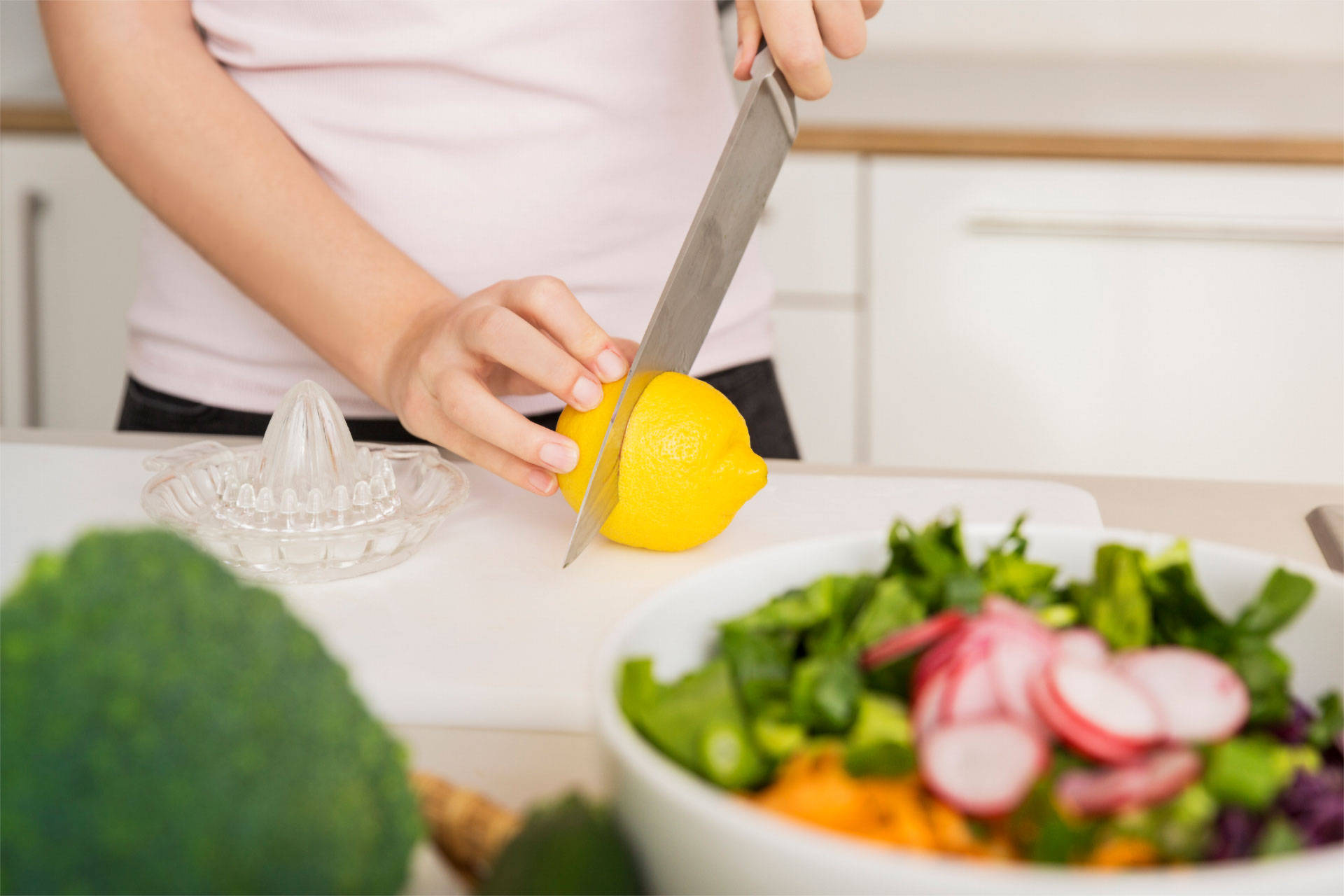 Person schneidet eine Zitrone auf einem Schneidebrett, daneben eine Presse und eine Salatschüssel, mit frischem Gemüse.