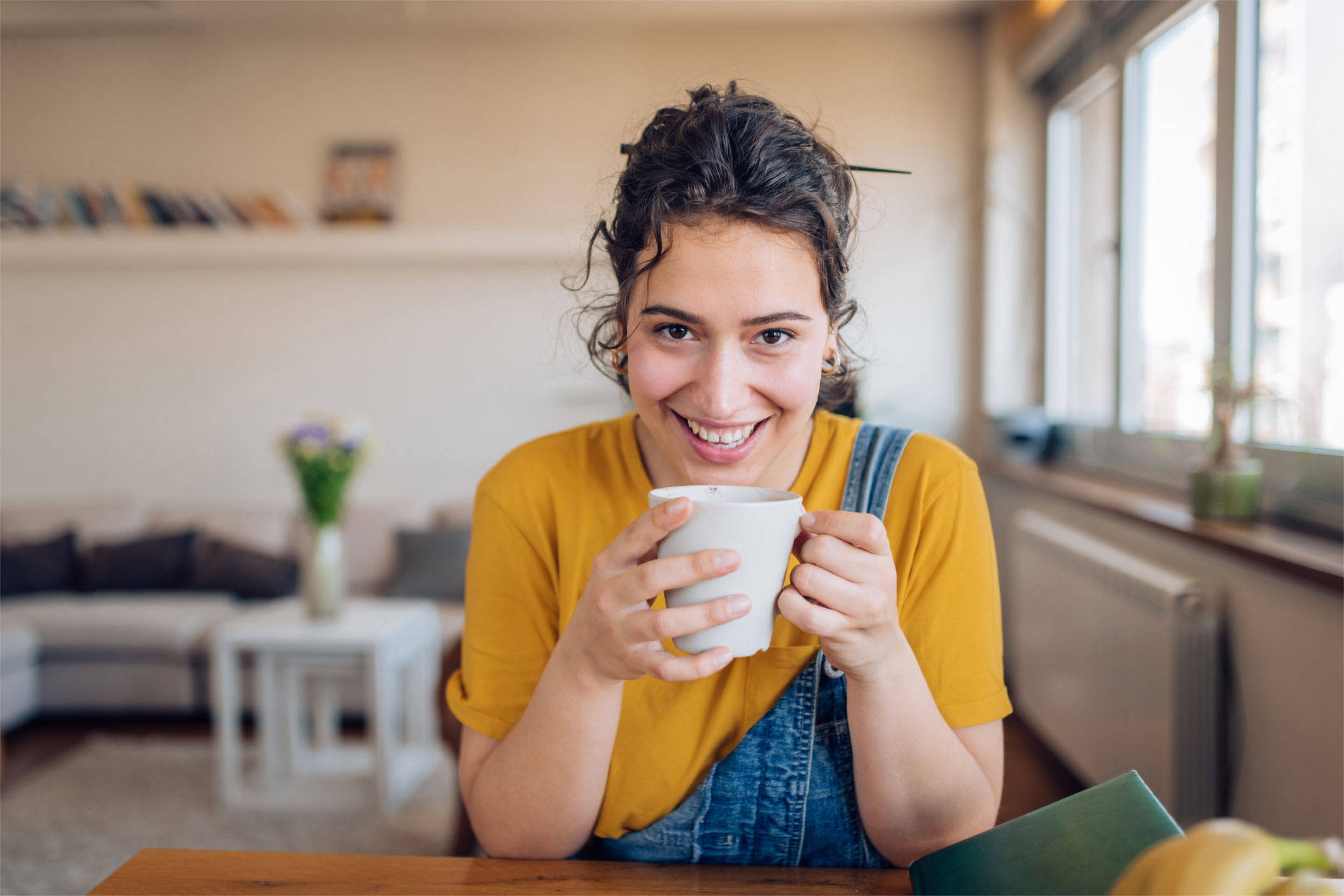 Junge Frau in gelbem Shirt und Latzhose hält eine Tasse und lächelt in die Kamera.