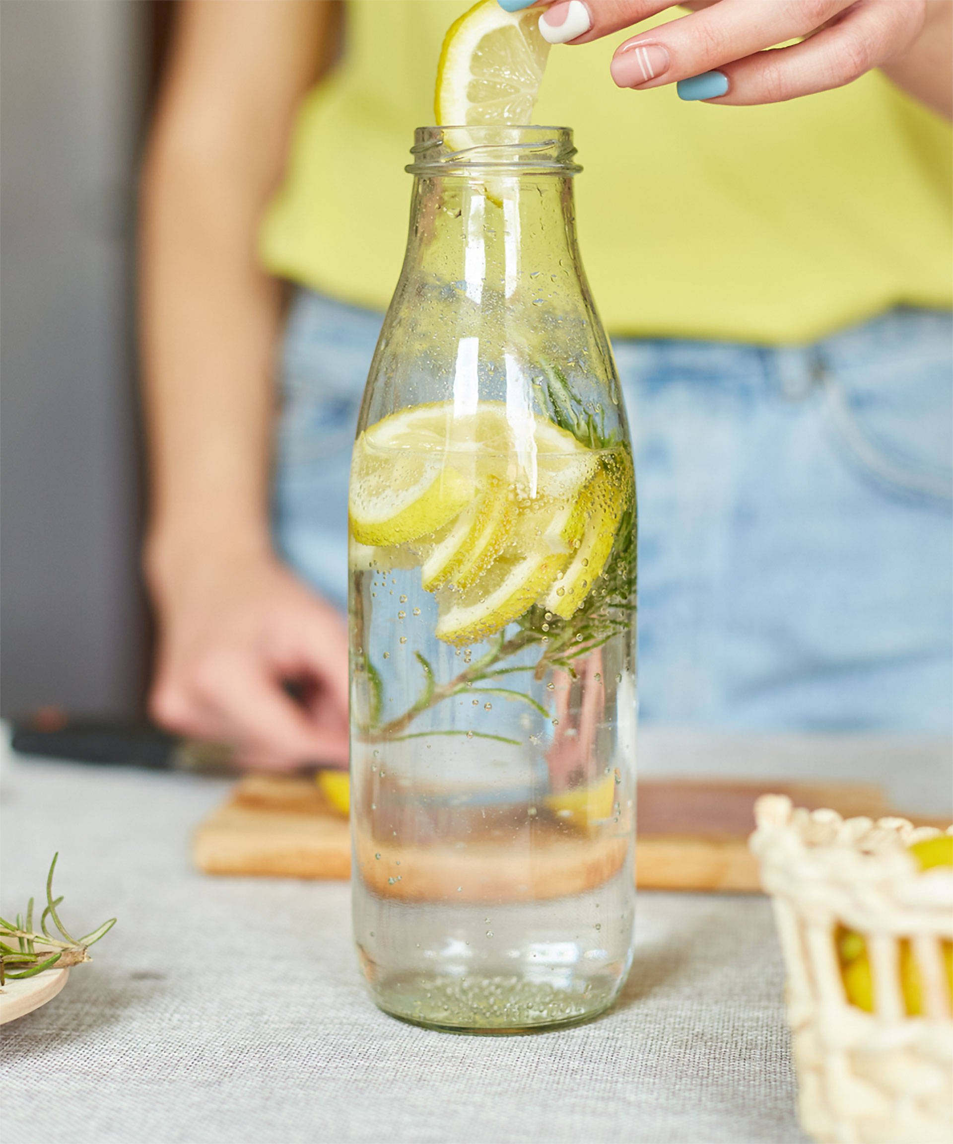 Person fügt Zitronenscheibe in eine Flasche Wasser mit Rosmarin, daneben Zitronen und ein Korb. 