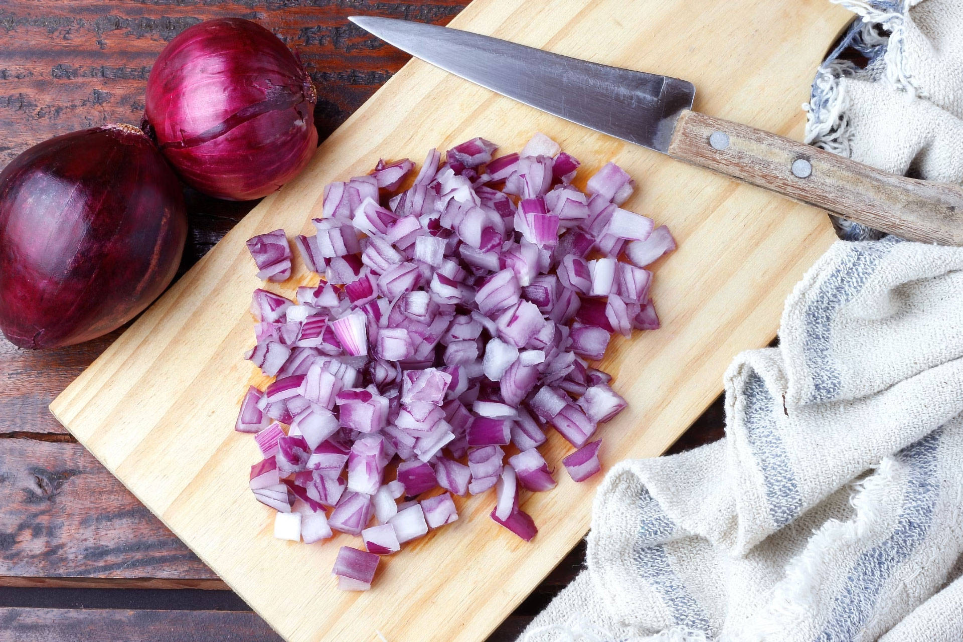 Gewürfelte rote Zwiebel auf einem Holzbrett neben einem Messer und zwei ganzen roten Zwiebeln.