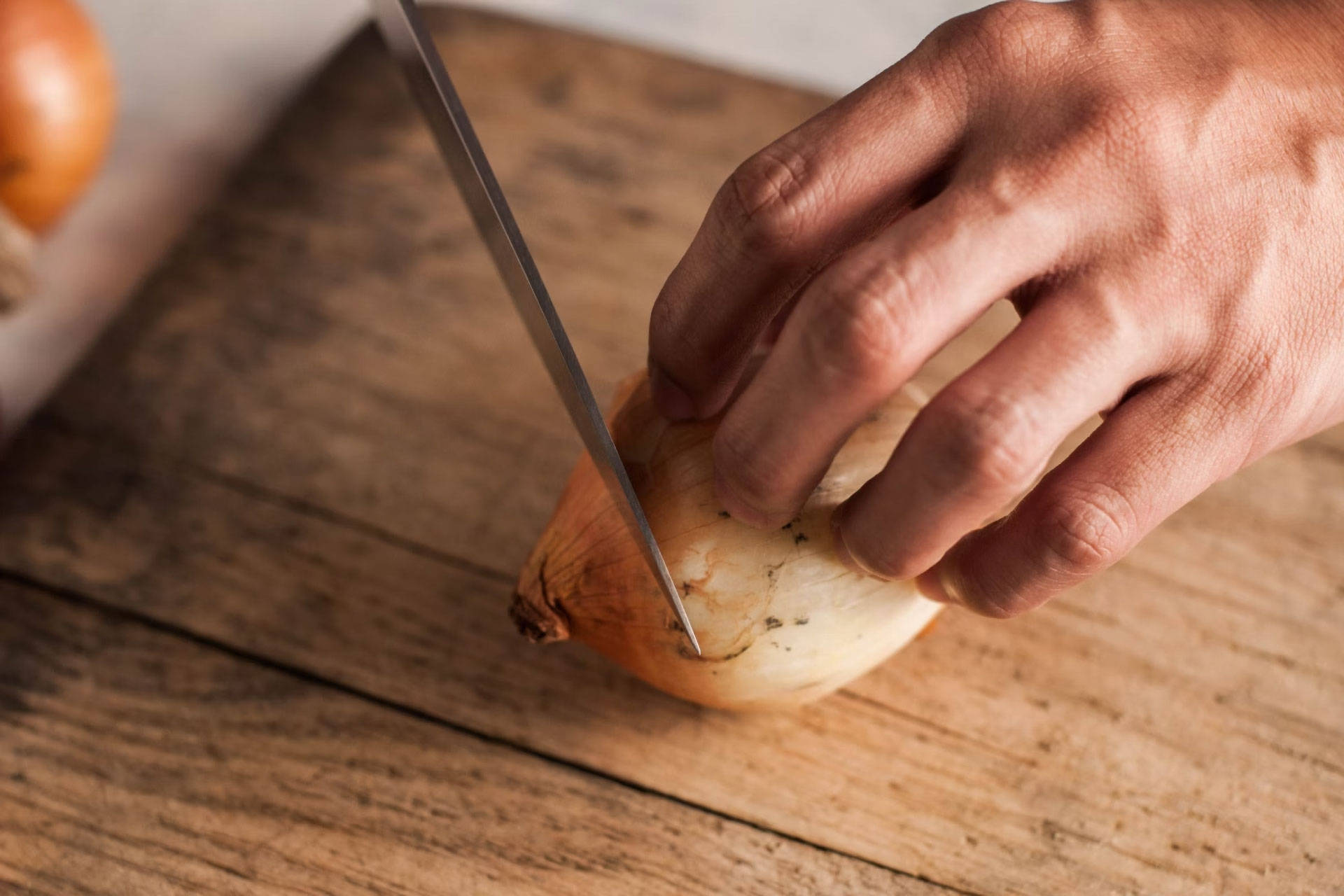 Person schneidet eine ungeschälte Zwiebel auf einem rustikalen Holzbrett mit einem scharfen Messer.