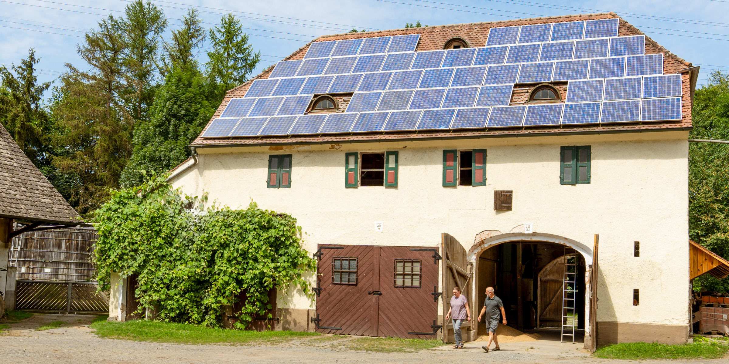 Ein Bauernhofgebäude mit Solarpanels auf dem Dach, umgeben von grüner Natur, zwei Personen gehen durch das Tor.