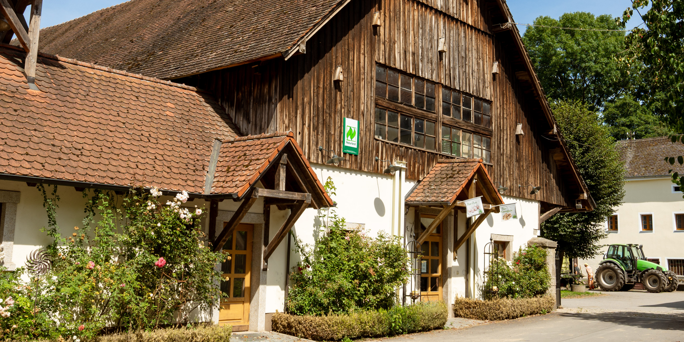 Ein traditionelles Bauernhofgebäude mit hölzernem Giebel, Ziegeldach und begrünten Fassaden, ein Traktor steht im Hof.