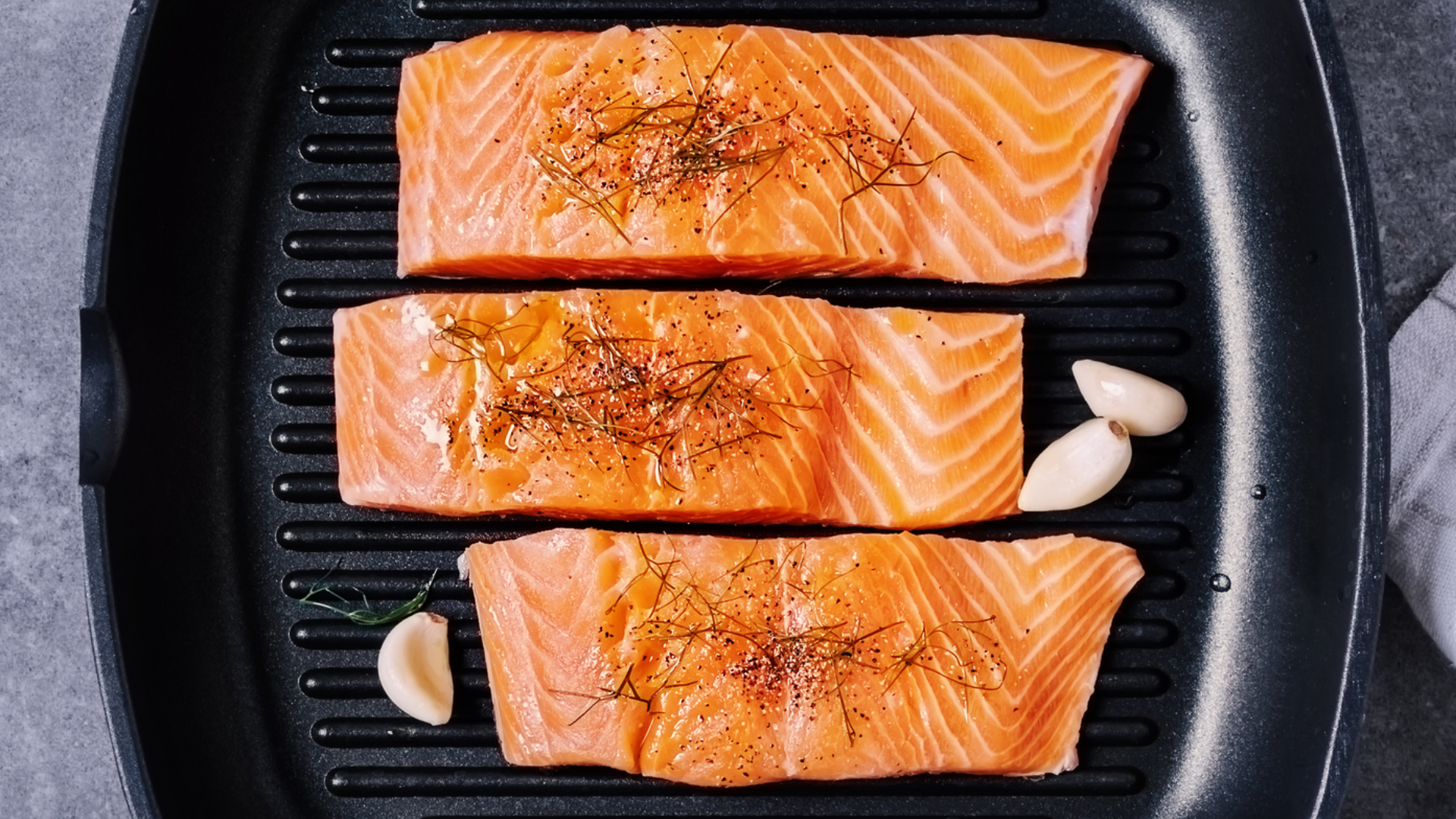 Drei Lachsfilets in Grillpfanne mit Knoblauch und Gewürzen.