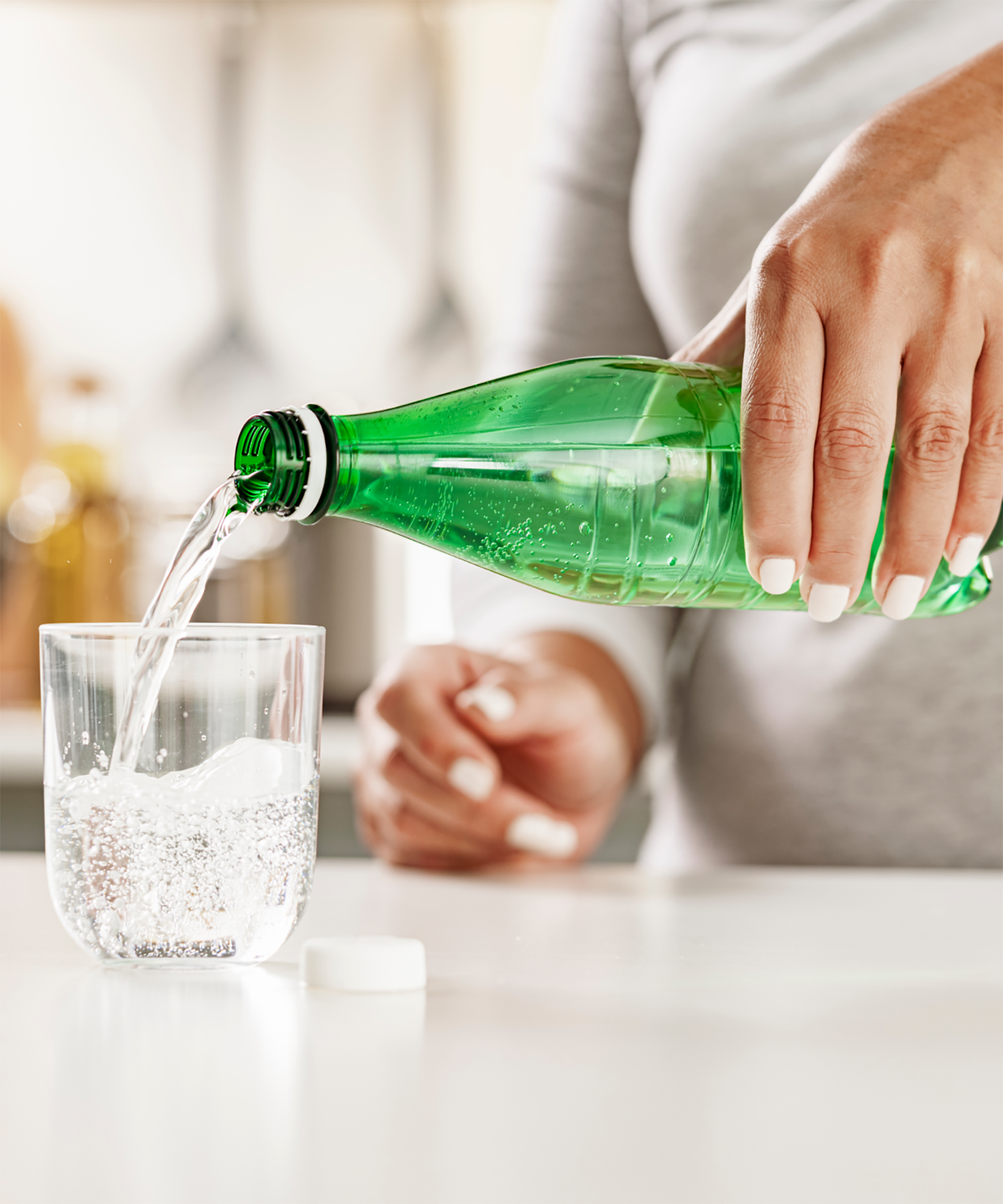 Person gießt Mineralwasser aus einer grünen PET-Flasche in ein Glas, das auf einer Küchenarbeitsplatte steht.