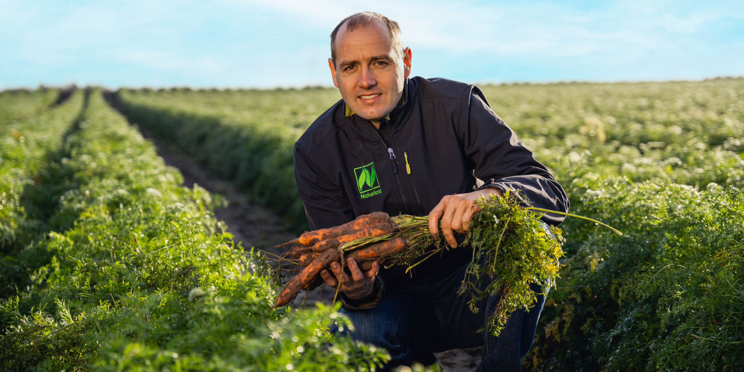 Biolandwirt Martin Schlereth hält frisch geerntete, erdige Karotten auf einem grünen Feld bei Sonnenschein.