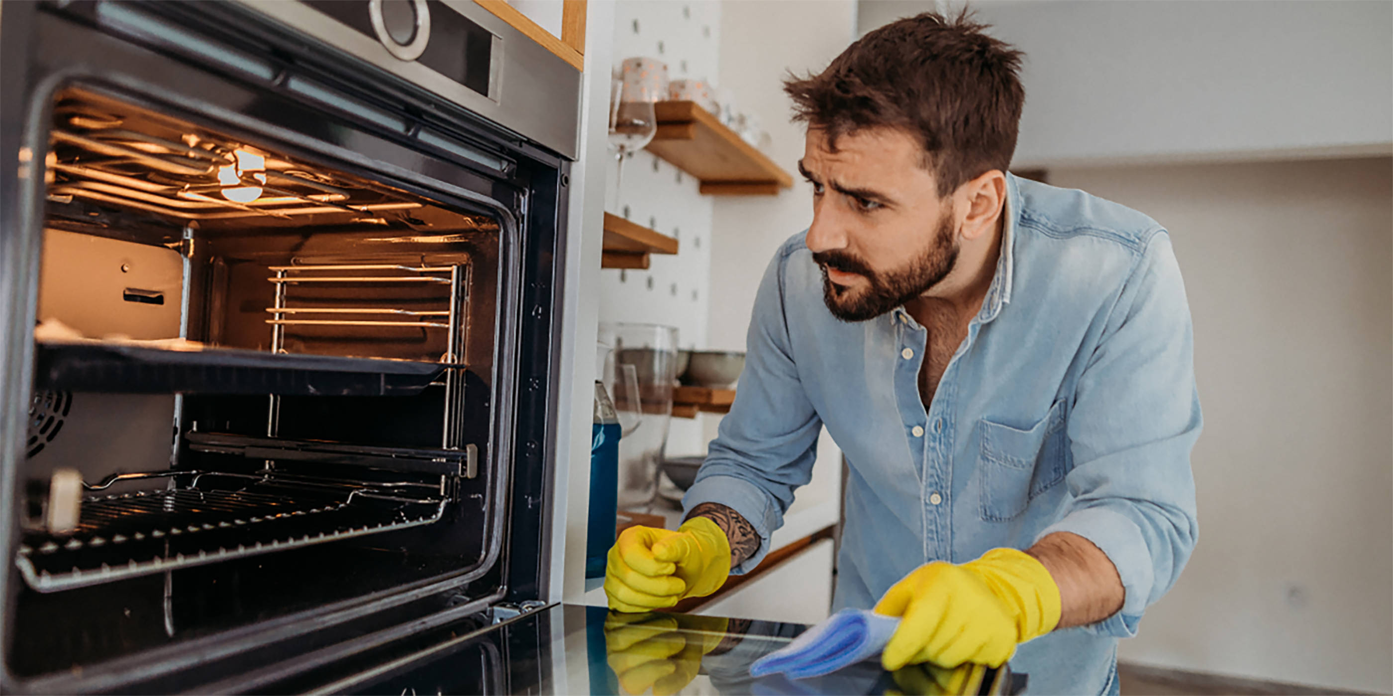 Ein Mann steht mit gelben Haushaltshandschuhen vor dem geöffneten Backofen, um ihn zu reinigen.