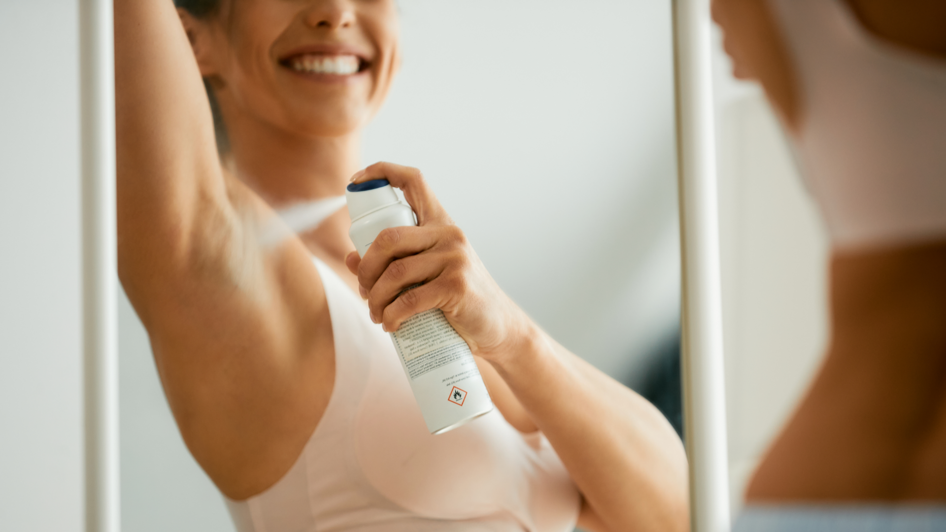 Frau sprüht Deodorant unter den Arm vor Spiegel.